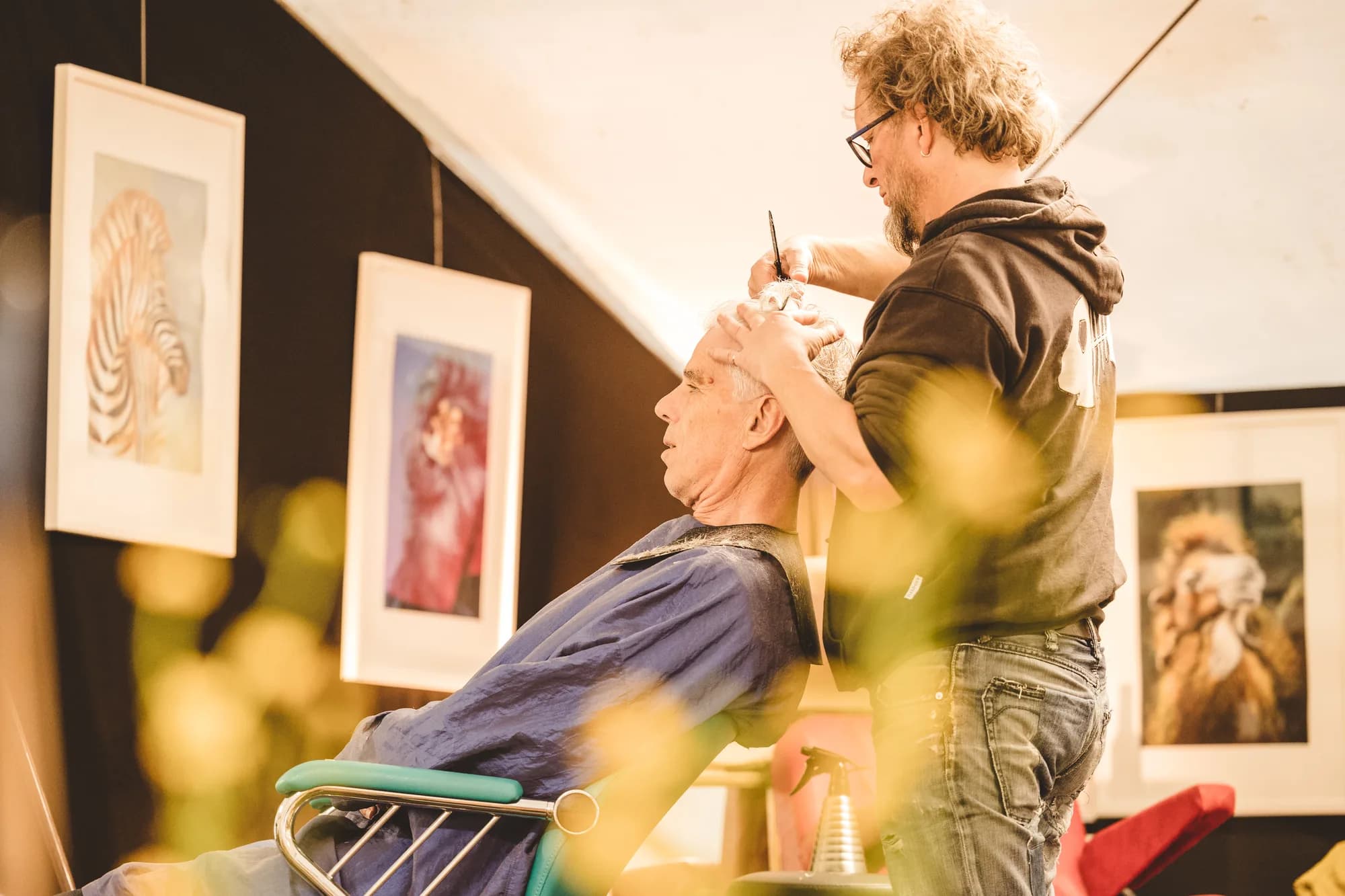 An artist gives a haircut to a seated man while framed animal portraits hang on the wall behind them.