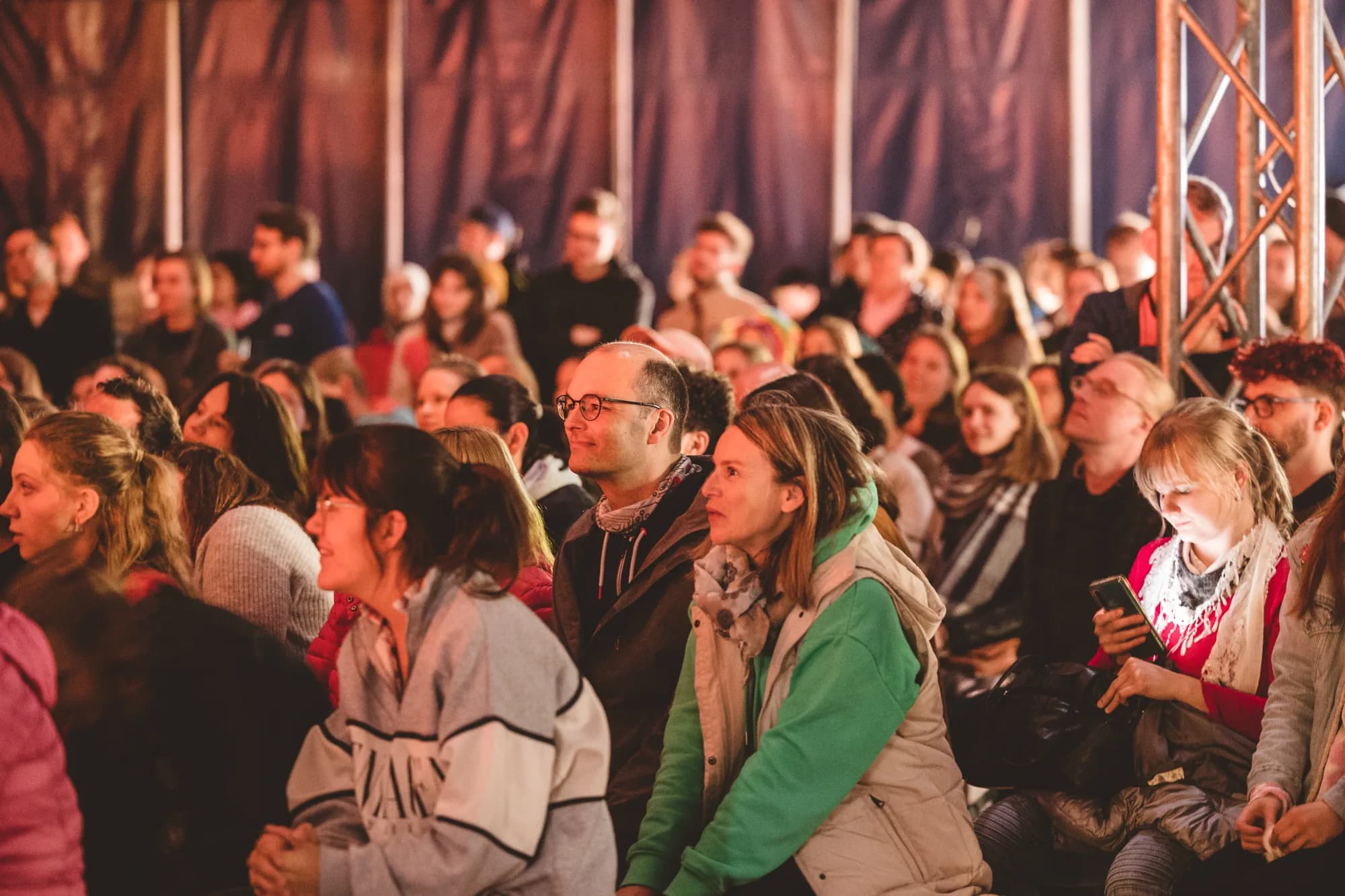 A seated audience of diverse adults watches a performance attentively inside a dimly lit tent with warm lighting.