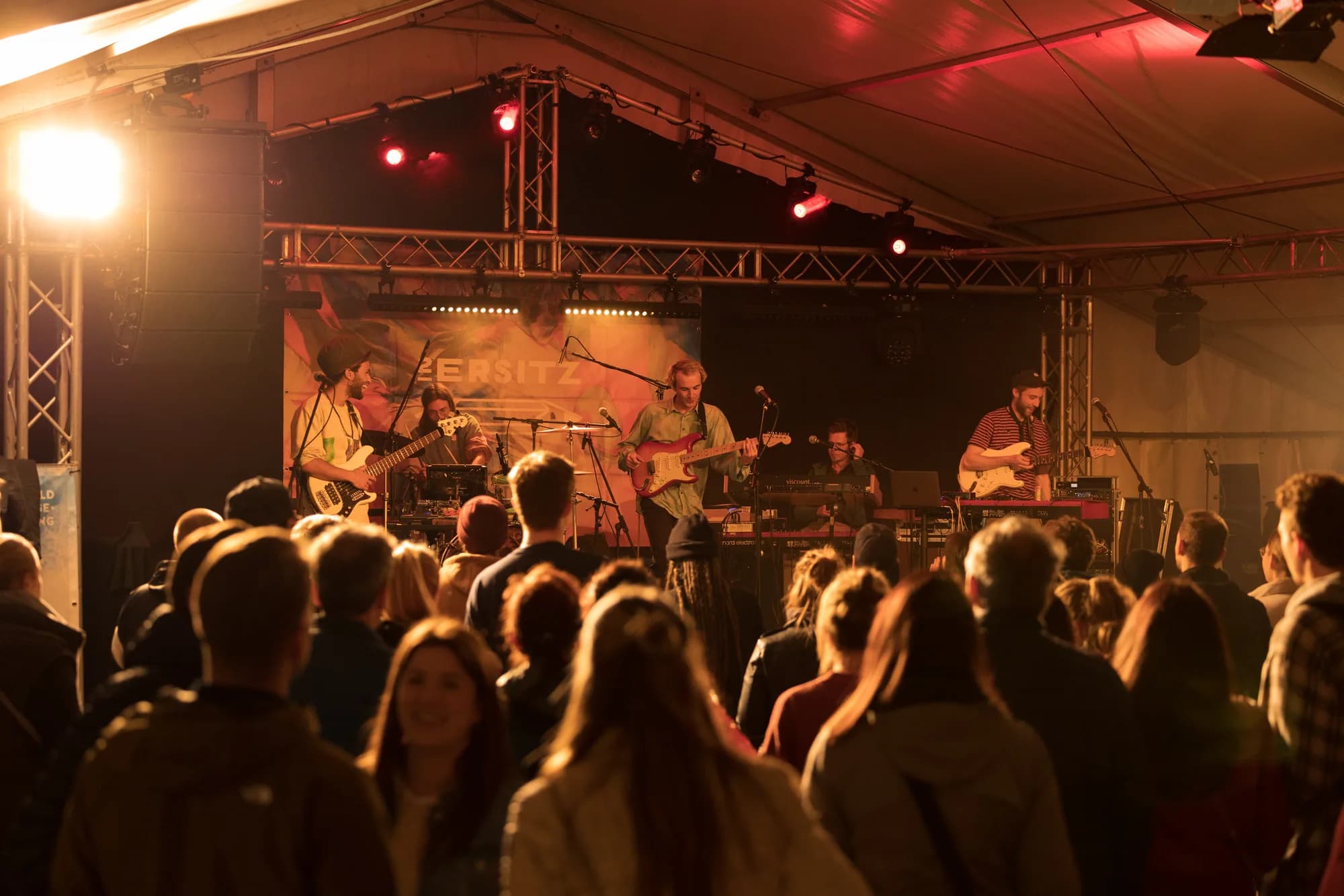 A folk band performs on a dimly lit stage inside a large tent while an audience watches from the front.