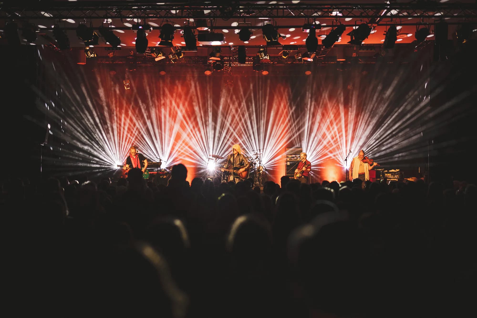 A four-piece band performs on an outdoor stage bathed in dramatic orange and white spotlights while a crowd watches in the foreground.