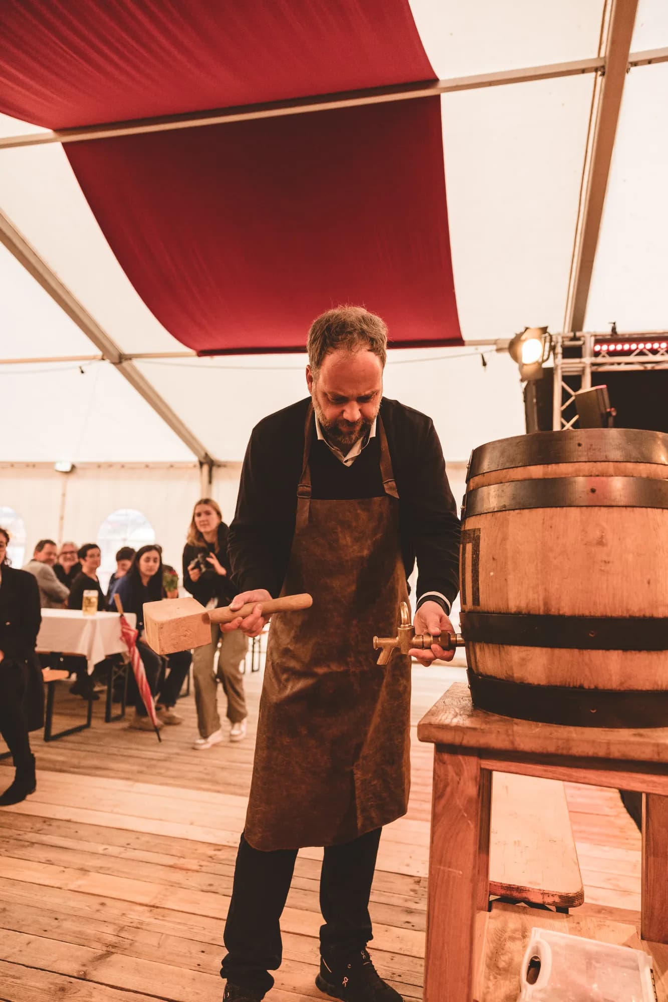 A man in a leather apron taps a wooden barrel with a mallet inside a white tent.