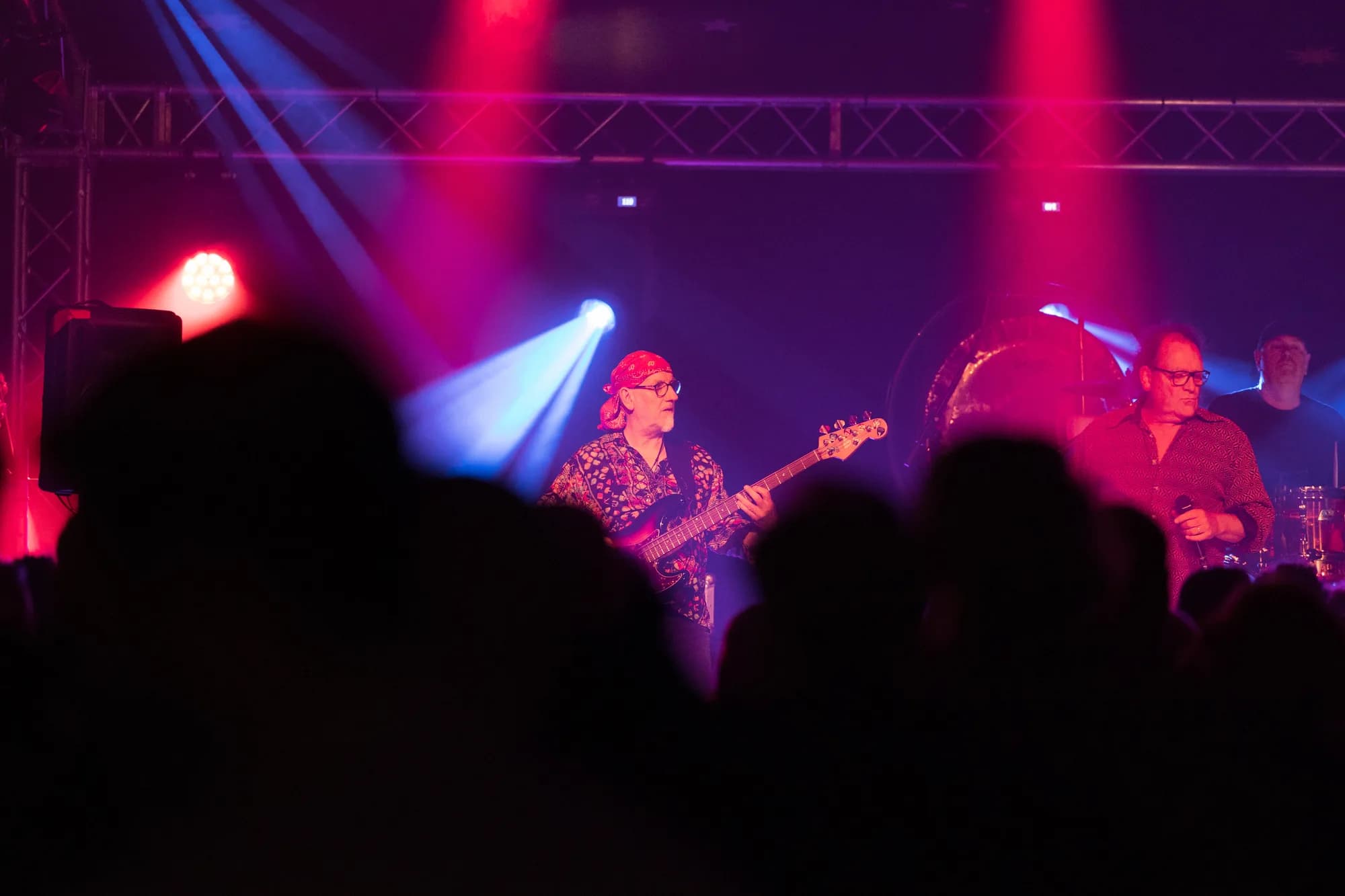 A bassist wearing a red bandana plays on stage illuminated by pink and blue spotlights while the crowd watches.