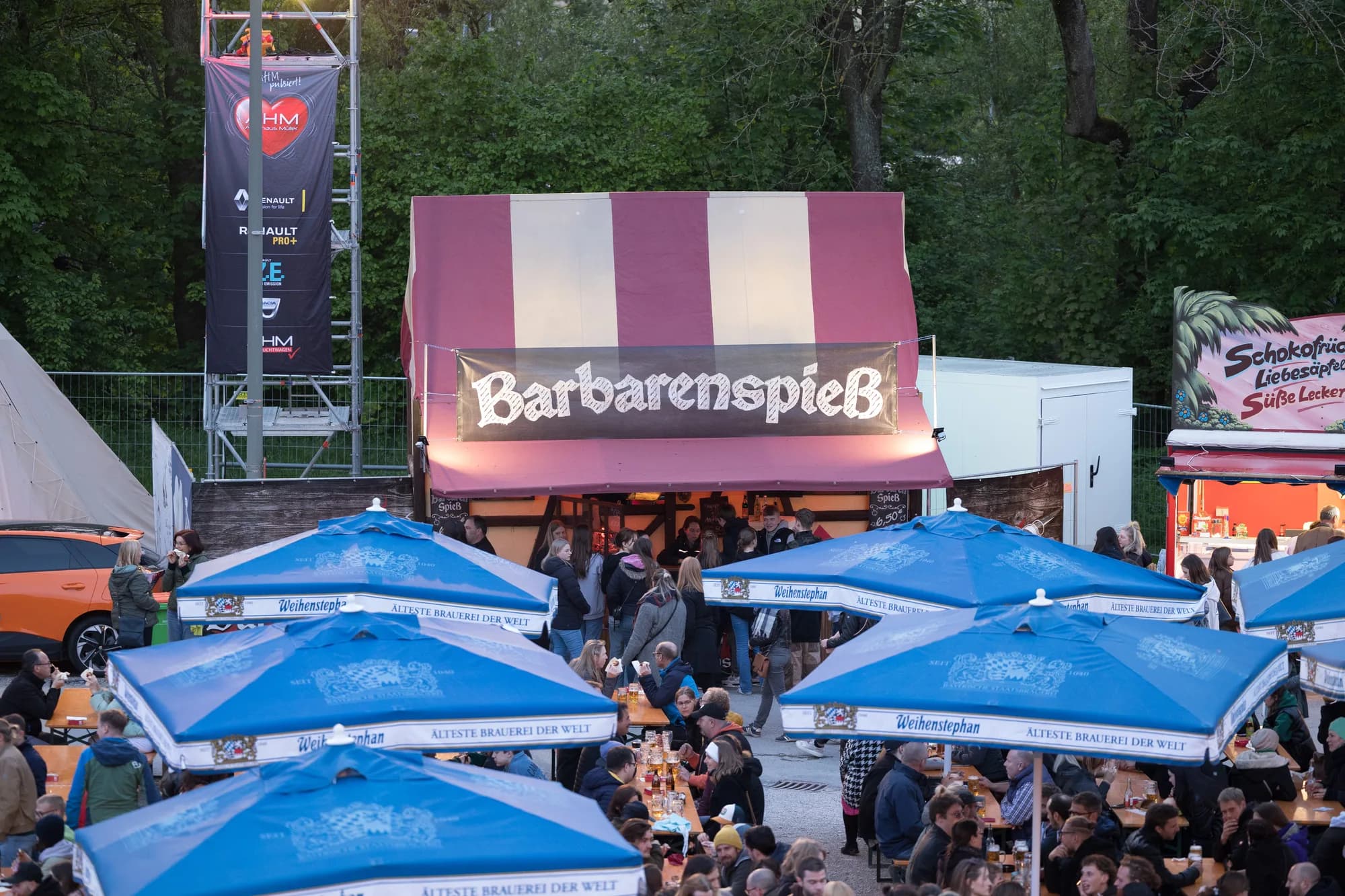 A busy beer garden at the Uferlos Festival with the Barbarenspieß food stall and blue umbrellas.