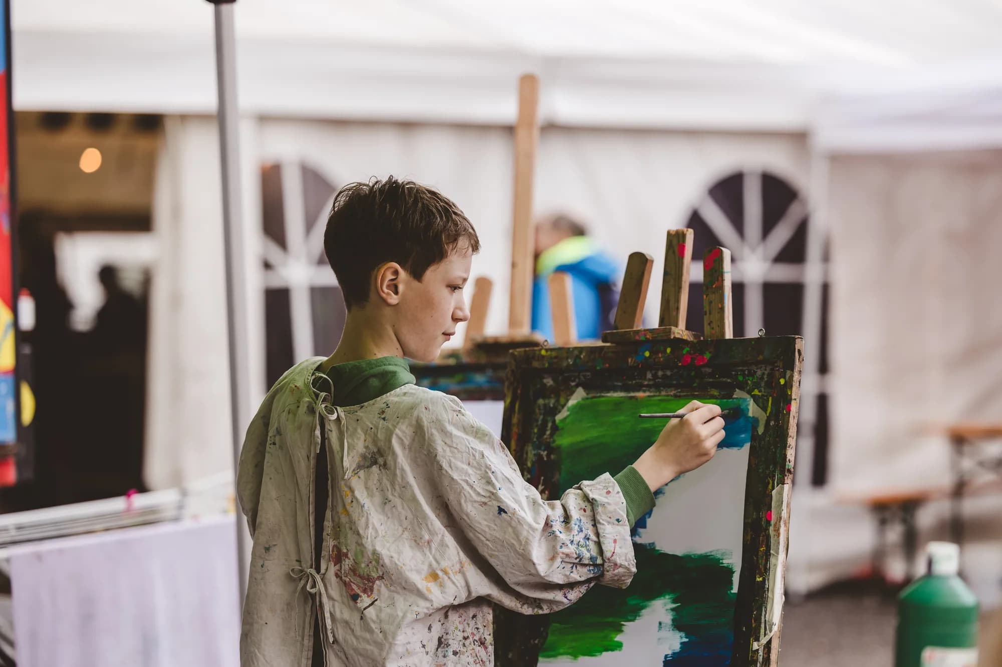 A young boy wearing a paint-splattered smock focuses intently on painting a green landscape on an easel.