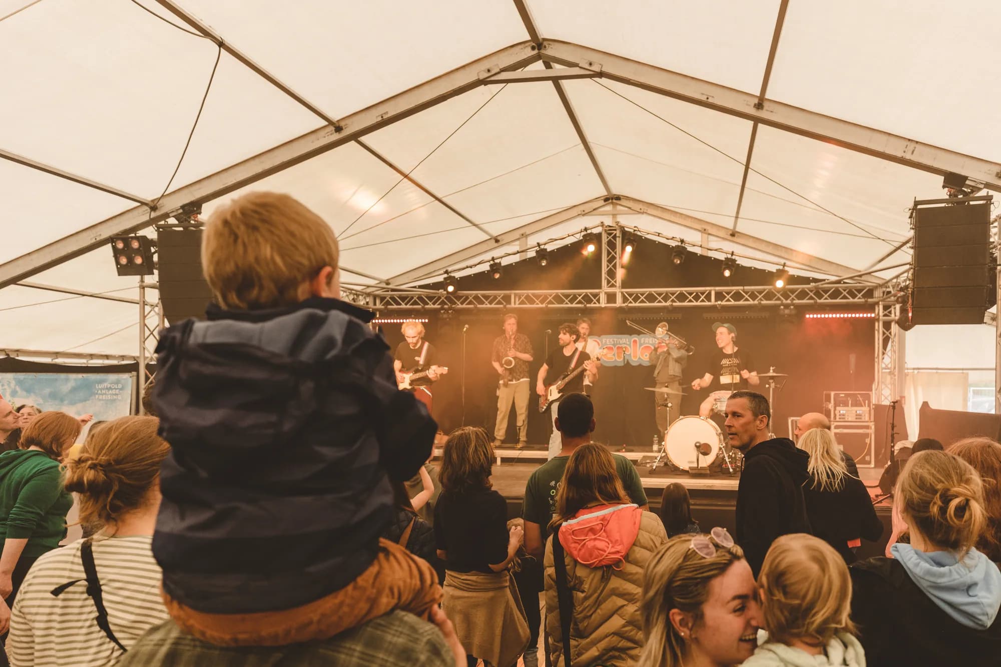 A brass band performs on stage inside a large white tent while a crowd watches, including a child sitting on an adult's shoulders.