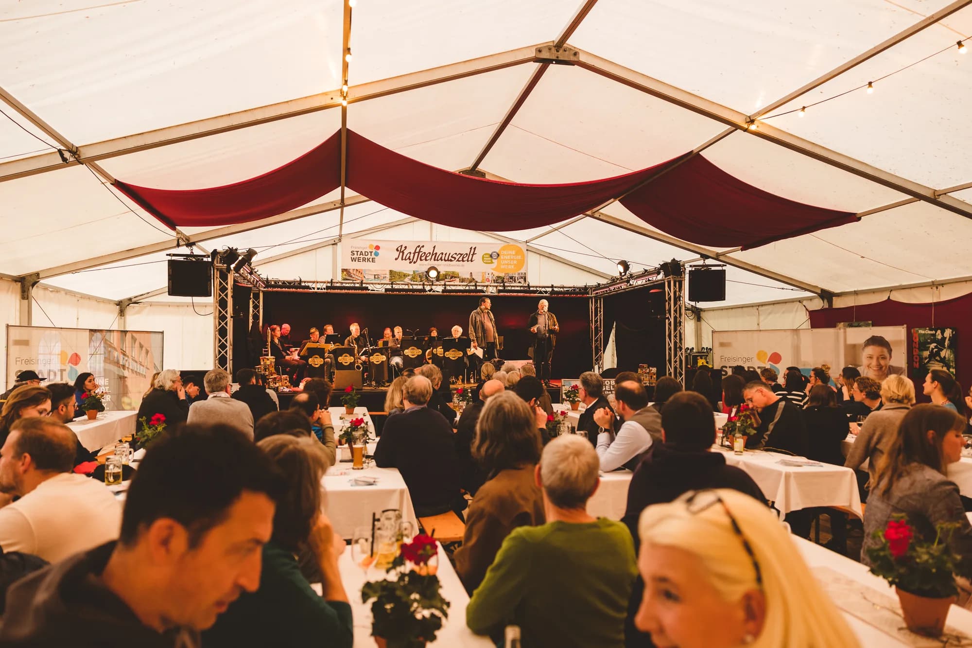 A brass band performs on stage inside the Kaffeehauszelt while an audience eats at long tables.