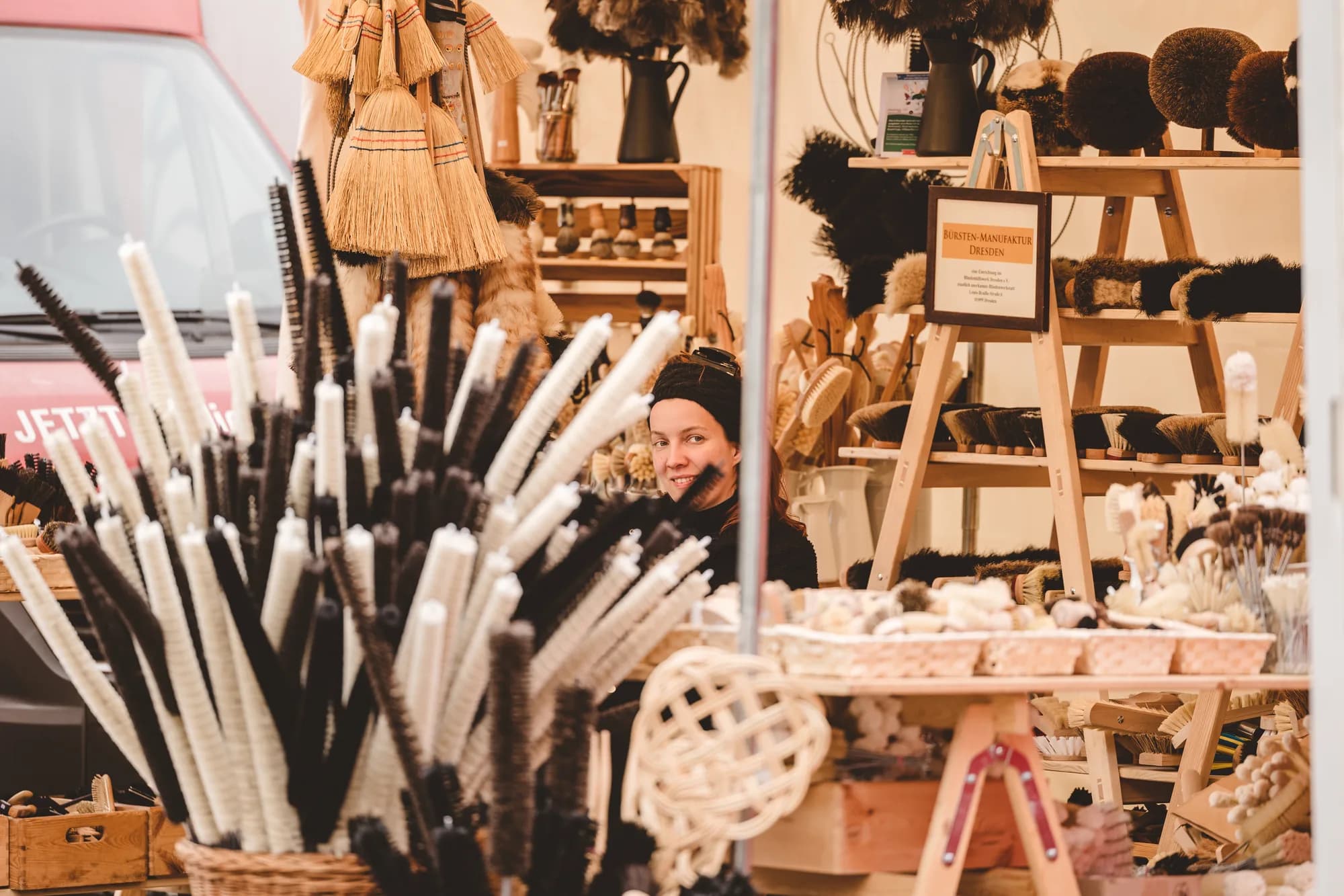 A vendor sits behind a wooden stall displaying handmade brooms and brushes at the Uferlos Festival market.
