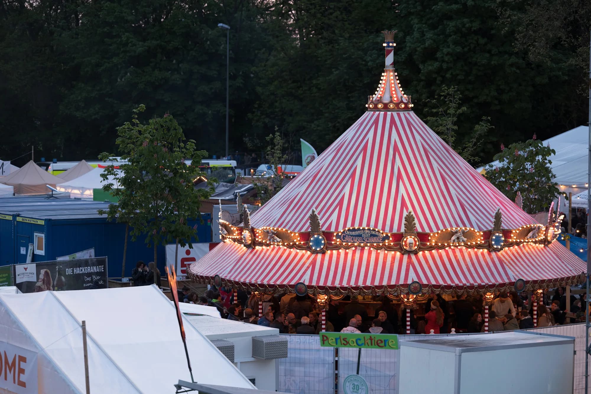 A red and white striped carousel illuminated with warm lights at dusk, surrounded by festival tents and trees.