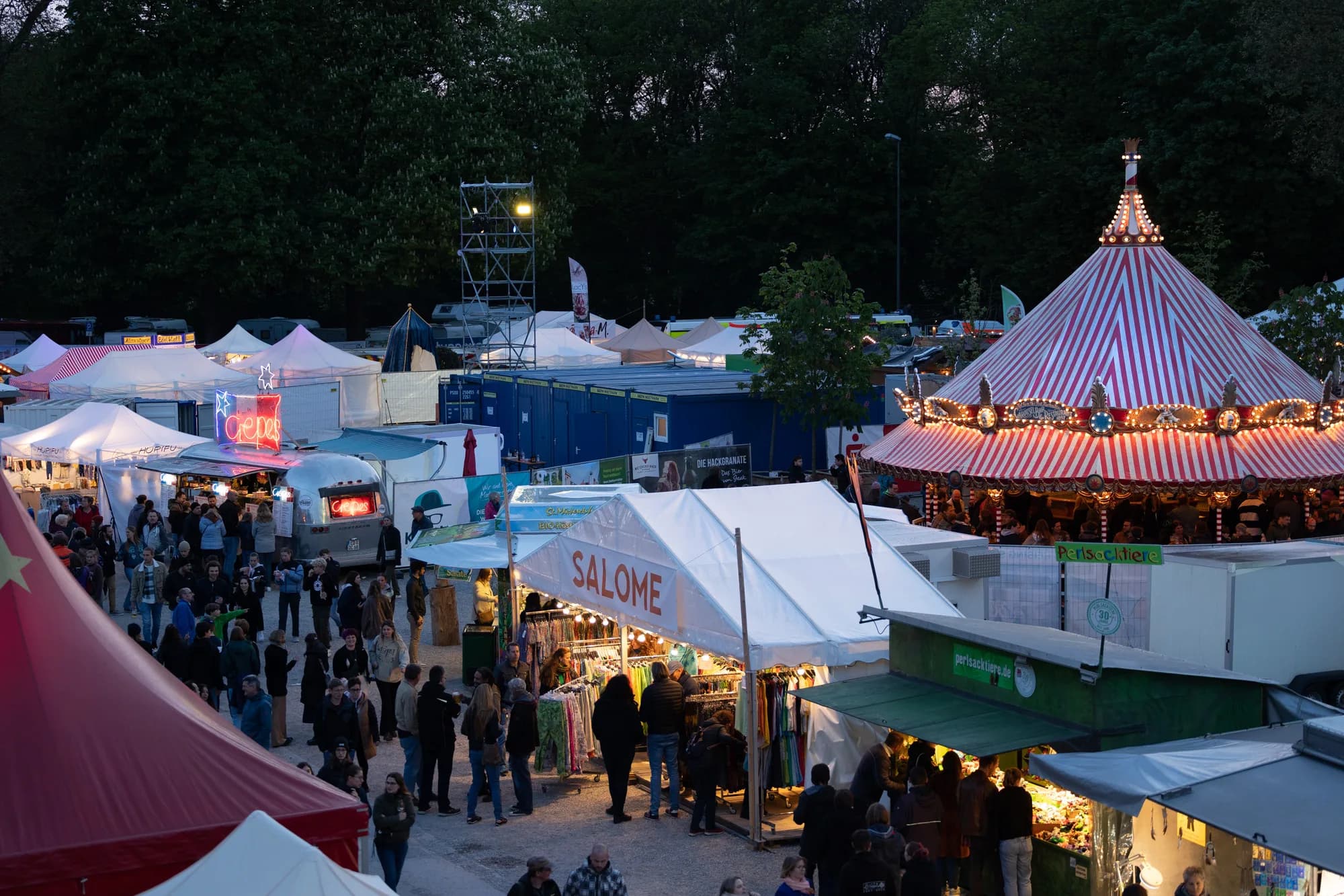 A high-angle view of the Uferlos festival market at dusk featuring a lit carousel and various vendor tents.