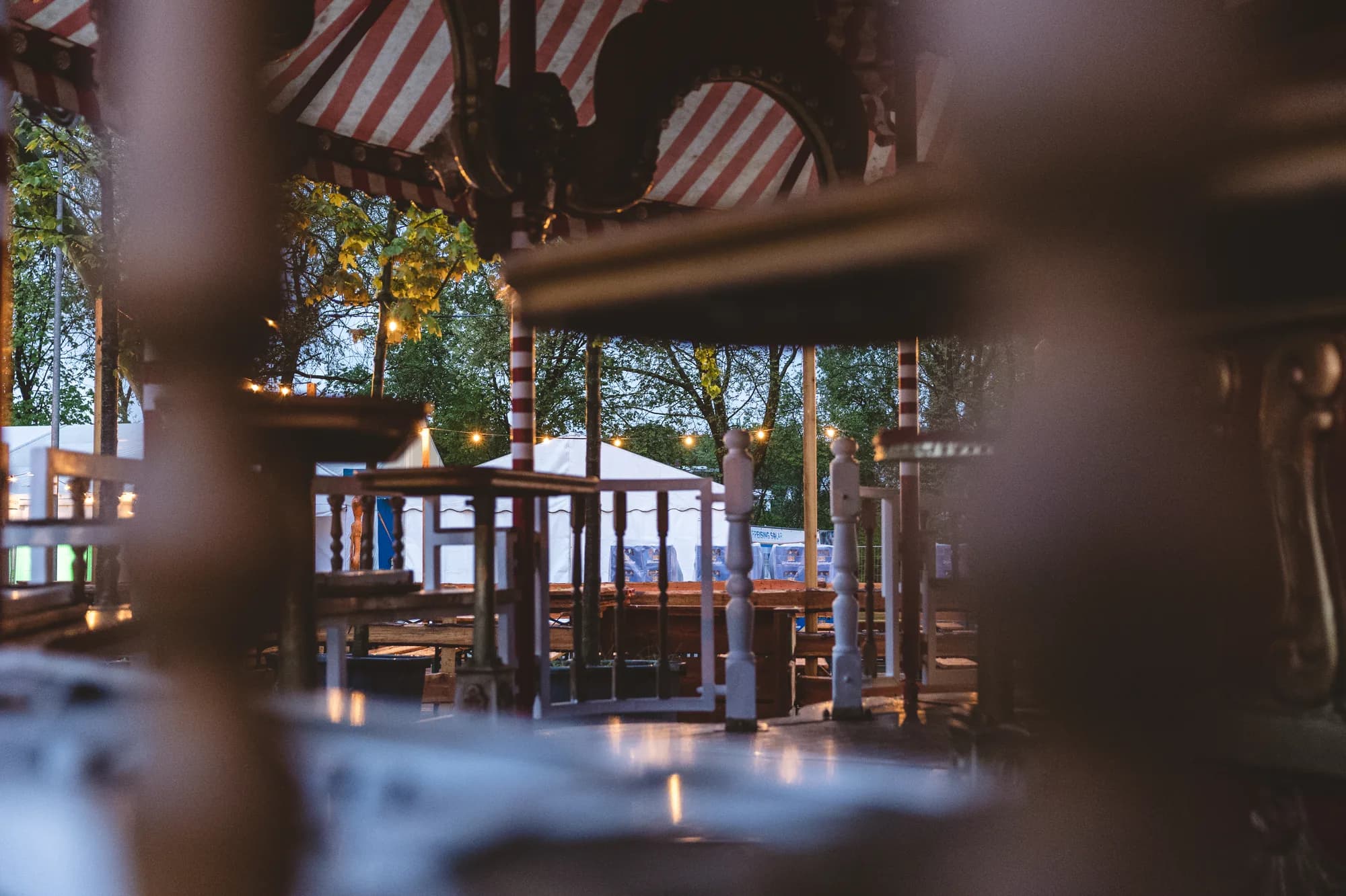 Close-up view of a wooden carousel structure with red and white striped awnings and warm string lights at dusk.
