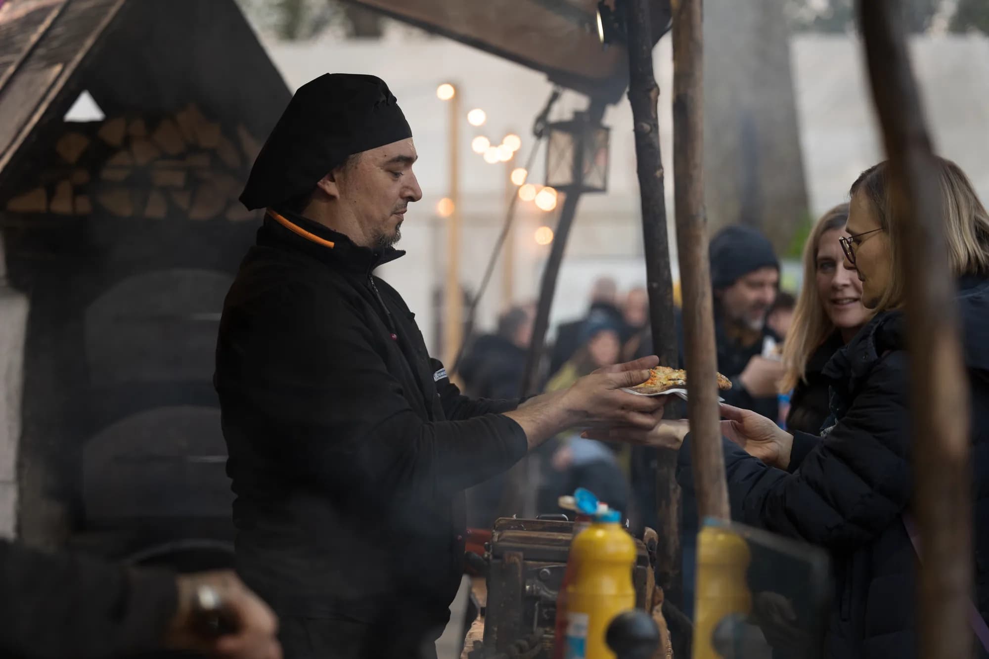A chef in a black hat hands a slice of pizza to a smiling customer at an outdoor food stall.