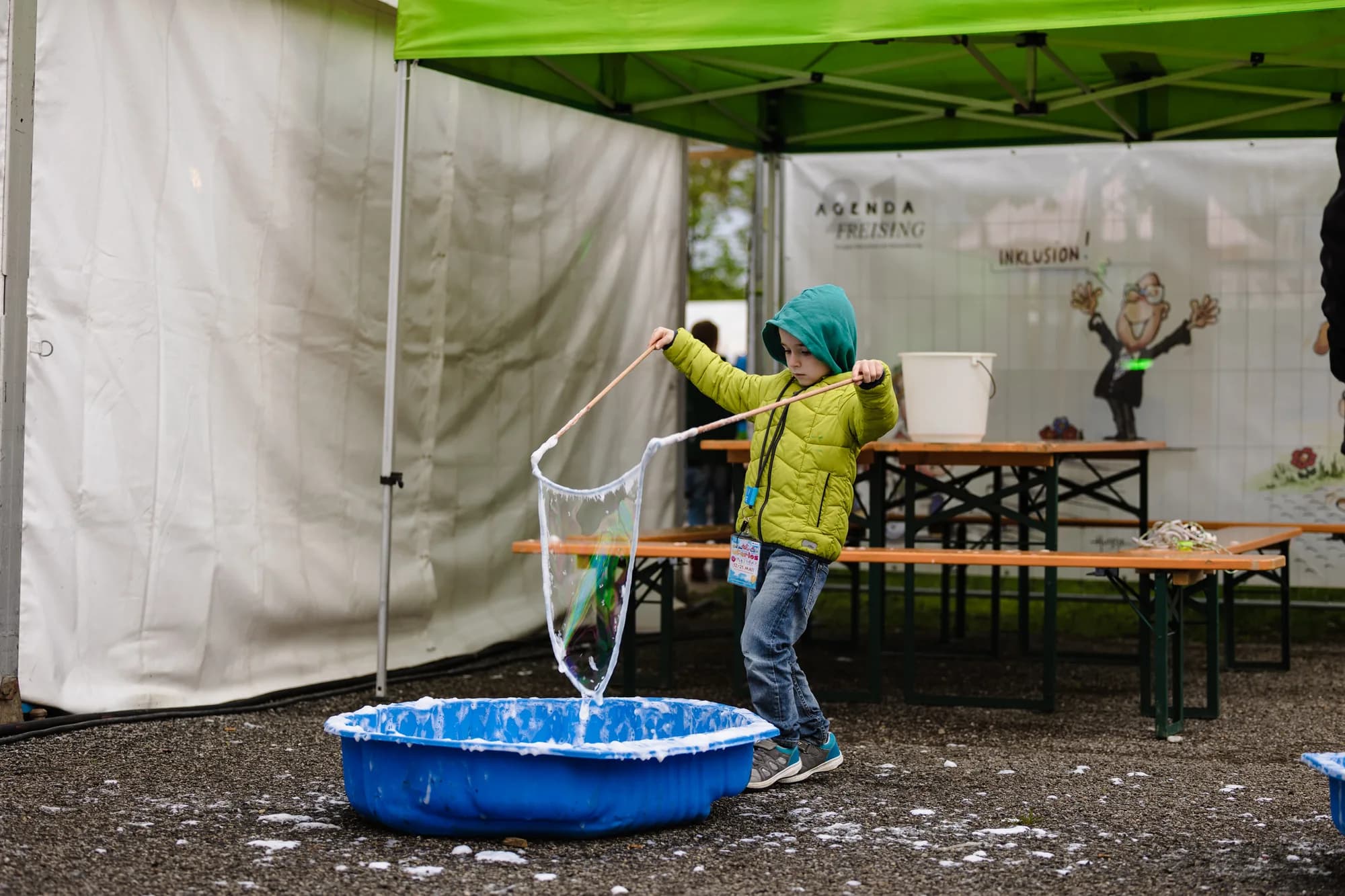 A young child in a green hooded jacket uses a large wand to create bubbles in a blue tub under a green festival tent.