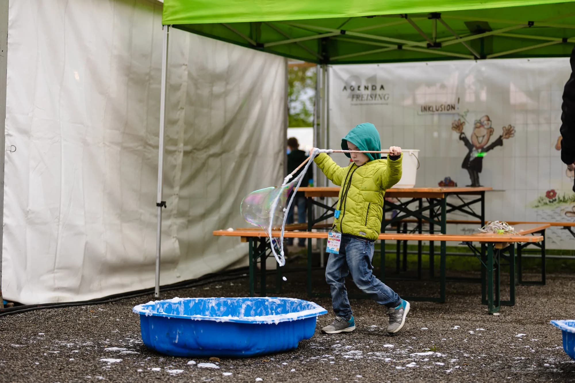 A young child in a yellow jacket creates giant bubbles in a blue tub at the children's tent.