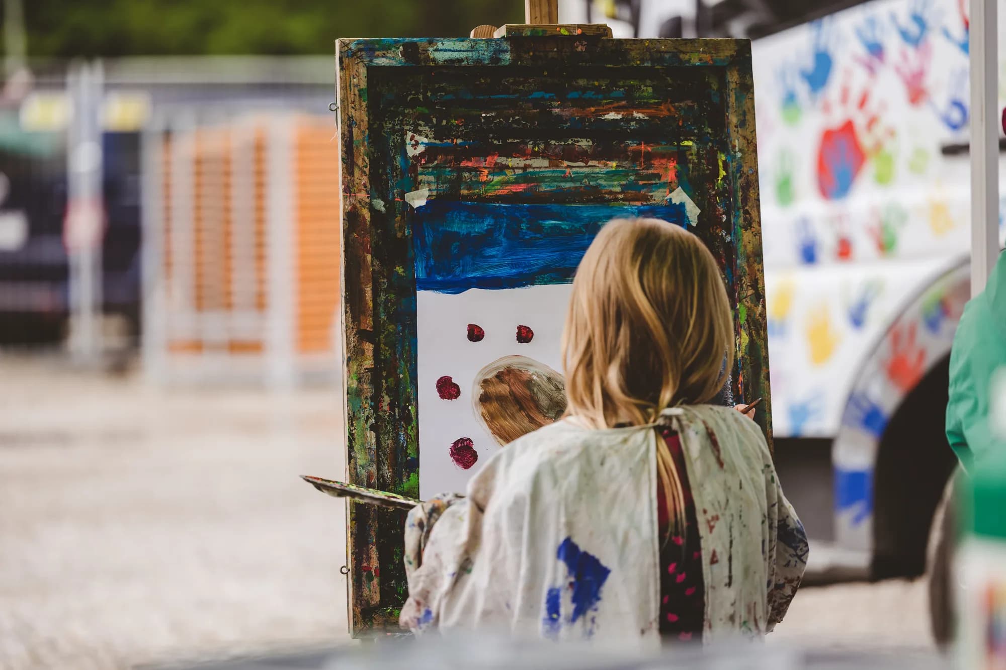 A young girl with blonde hair stands at an easel painting a picture of a snowman with red berries.