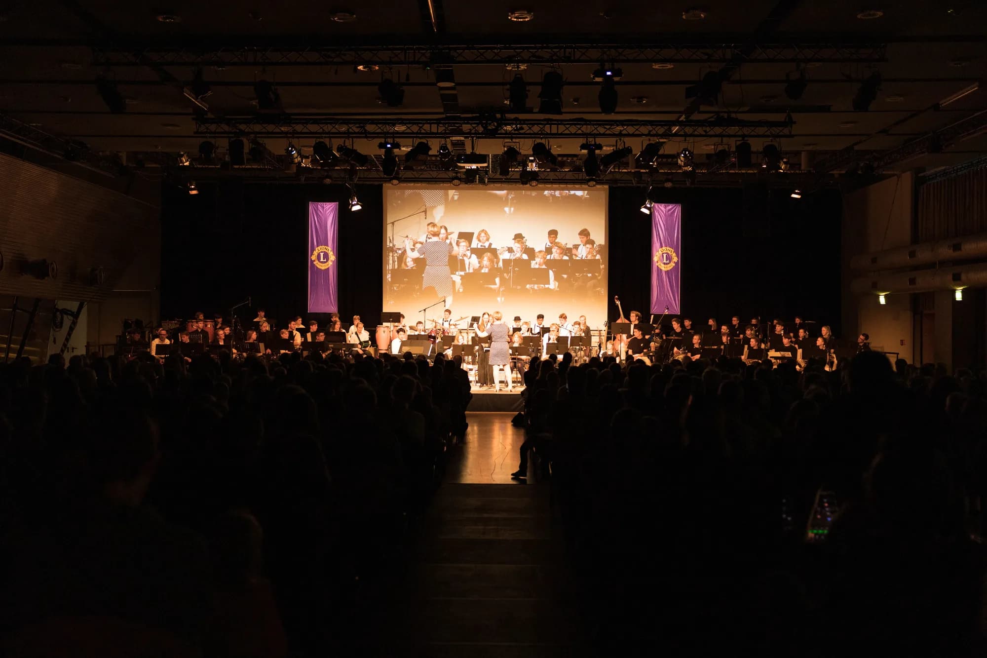 A large choir performs on a brightly lit stage with a conductor, projected on a screen behind them, while a dark audience watches from the floor.