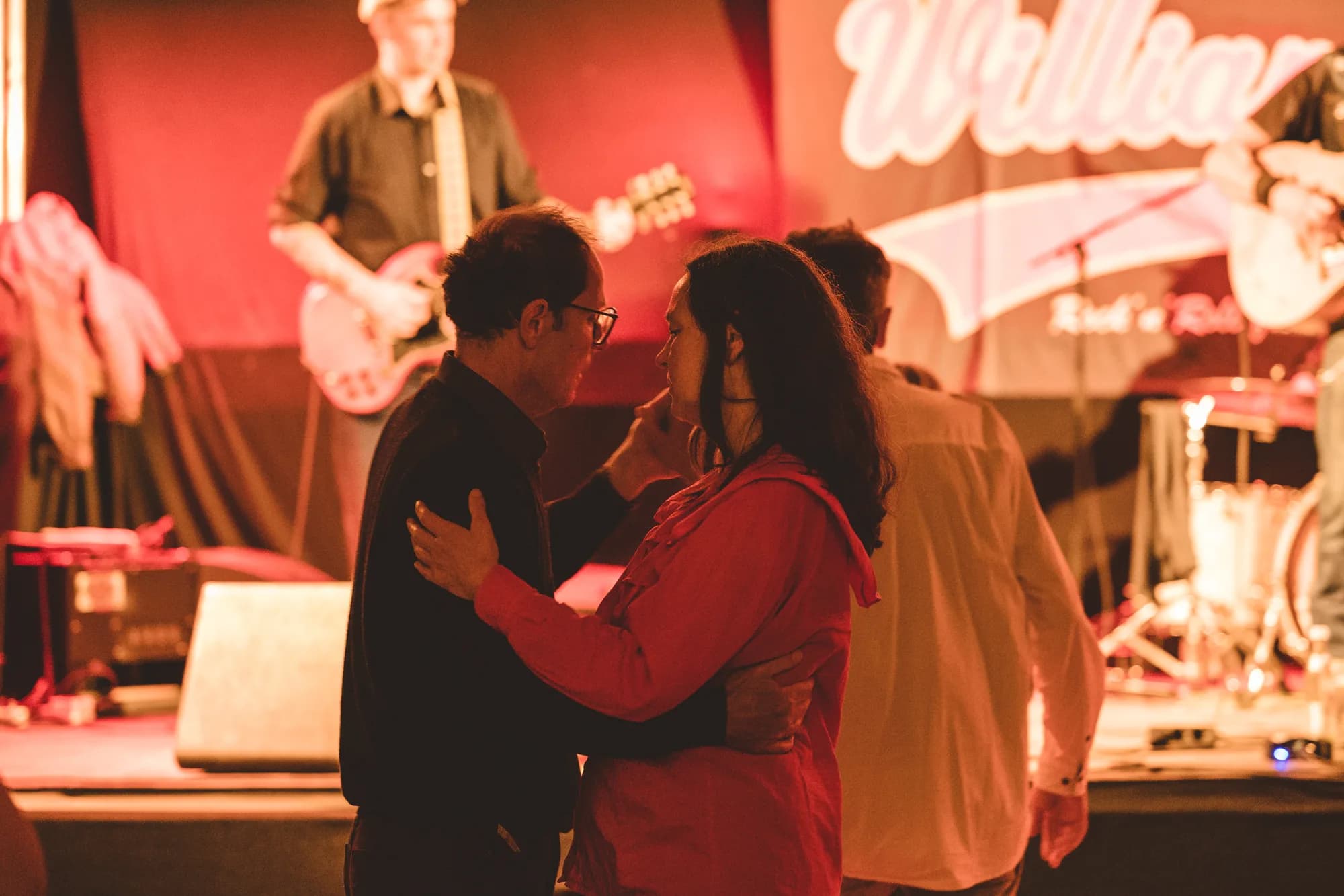 A couple dancing closely in the foreground while a rock band performs on a red-lit stage behind them.
