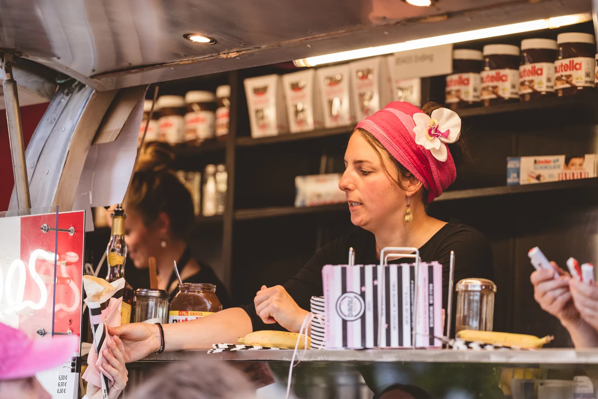A female vendor wearing a pink turban and flower serves a crepe to a customer at a festival food stall.