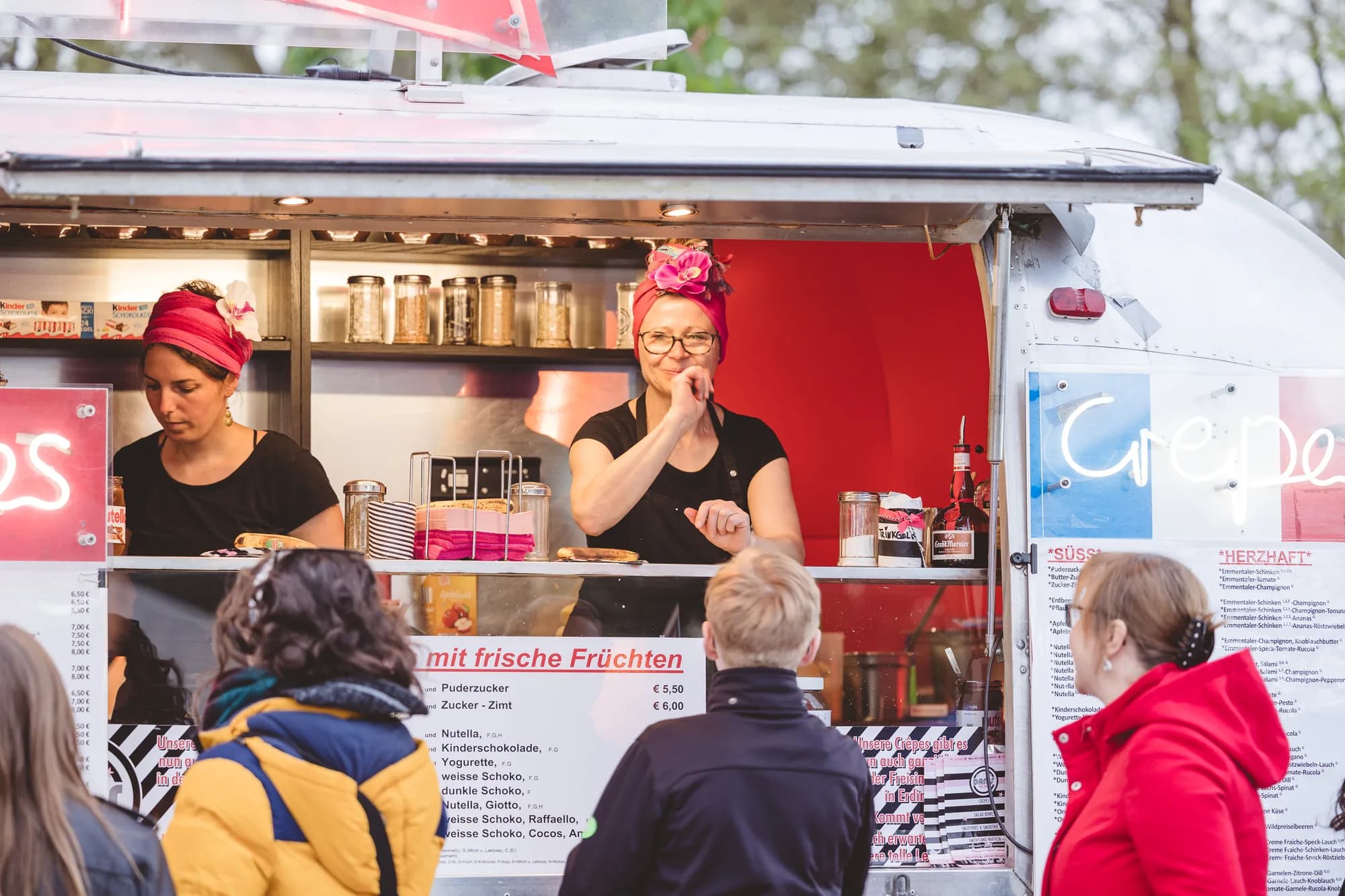 Two female vendors wearing red headscarves serve customers from a white food truck with a red interior.