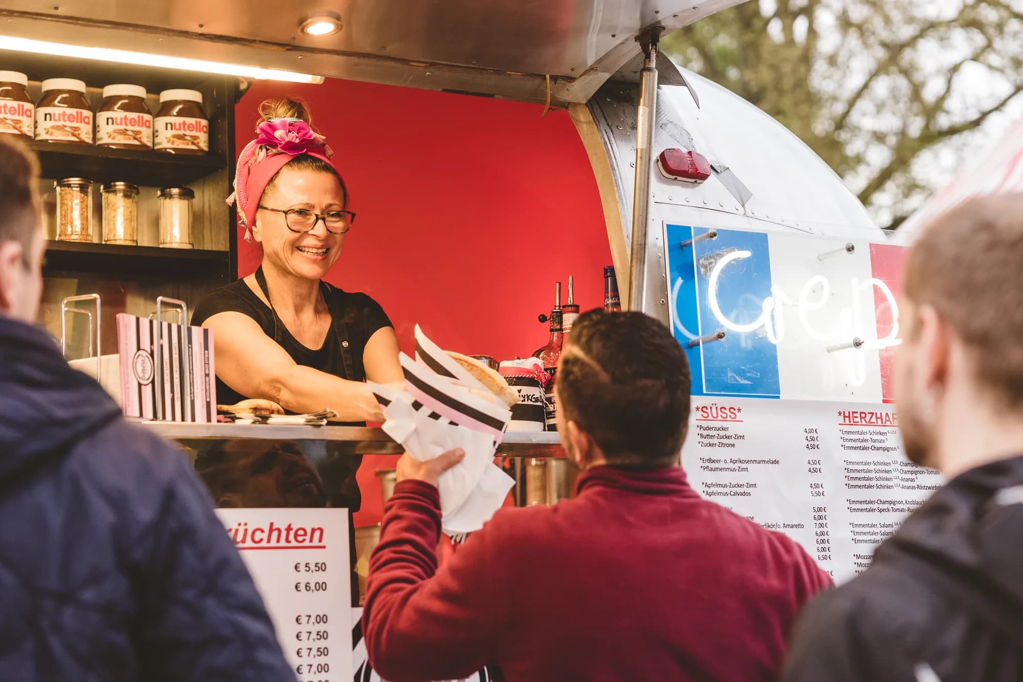 A smiling vendor with a pink headscarf serves a crepe to a customer at a food truck with a red interior.