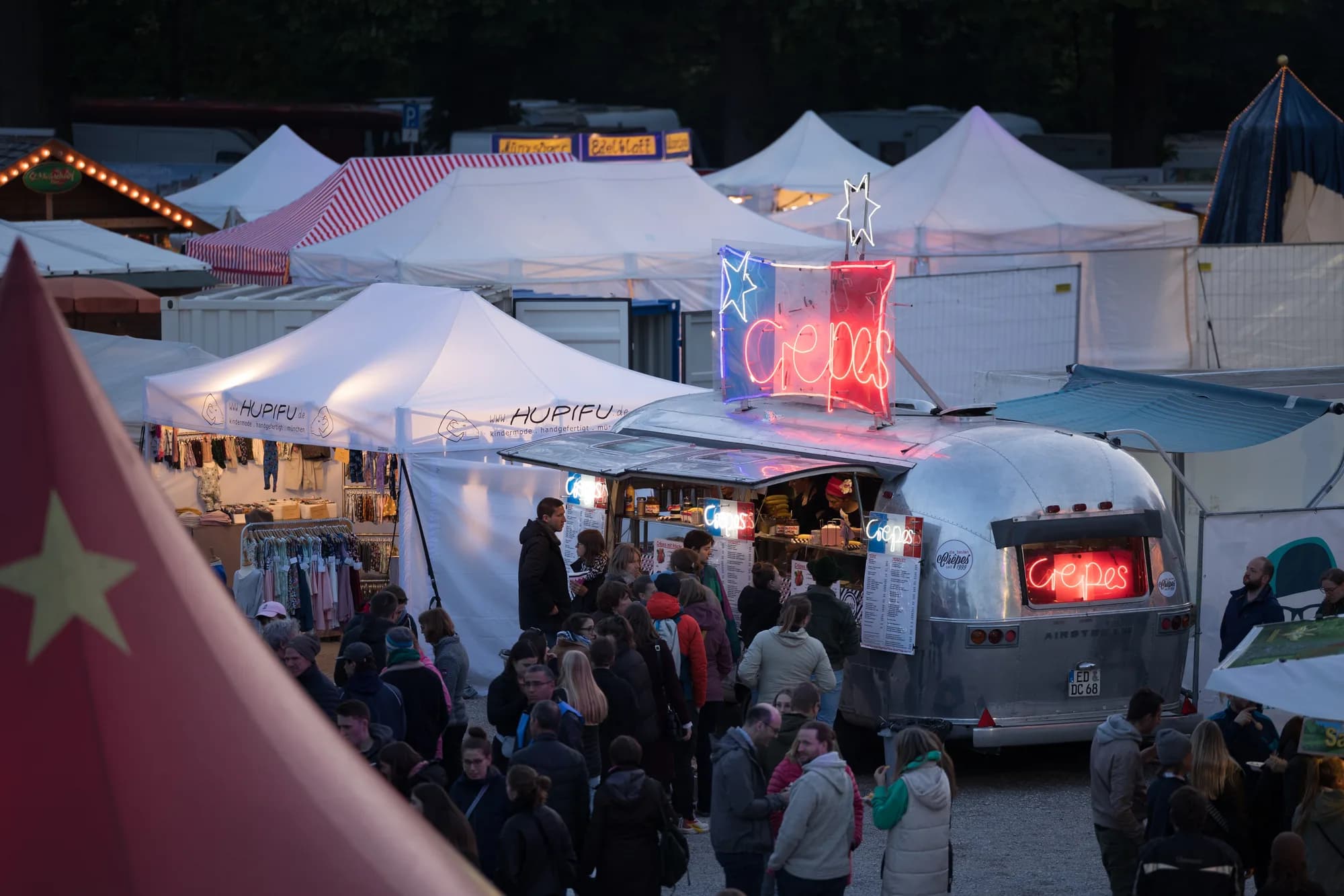 A silver Airstream food truck with neon 'crepes' signs serves a crowd of people in the festival market area at dusk.
