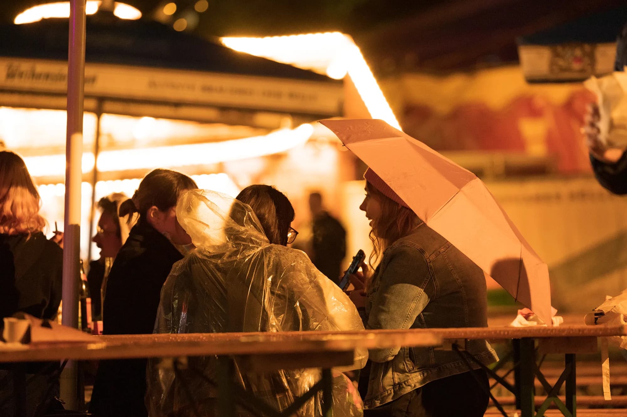 Festivalgoers huddle under an umbrella and plastic ponchos at a wooden table during a rainy night at the festival.