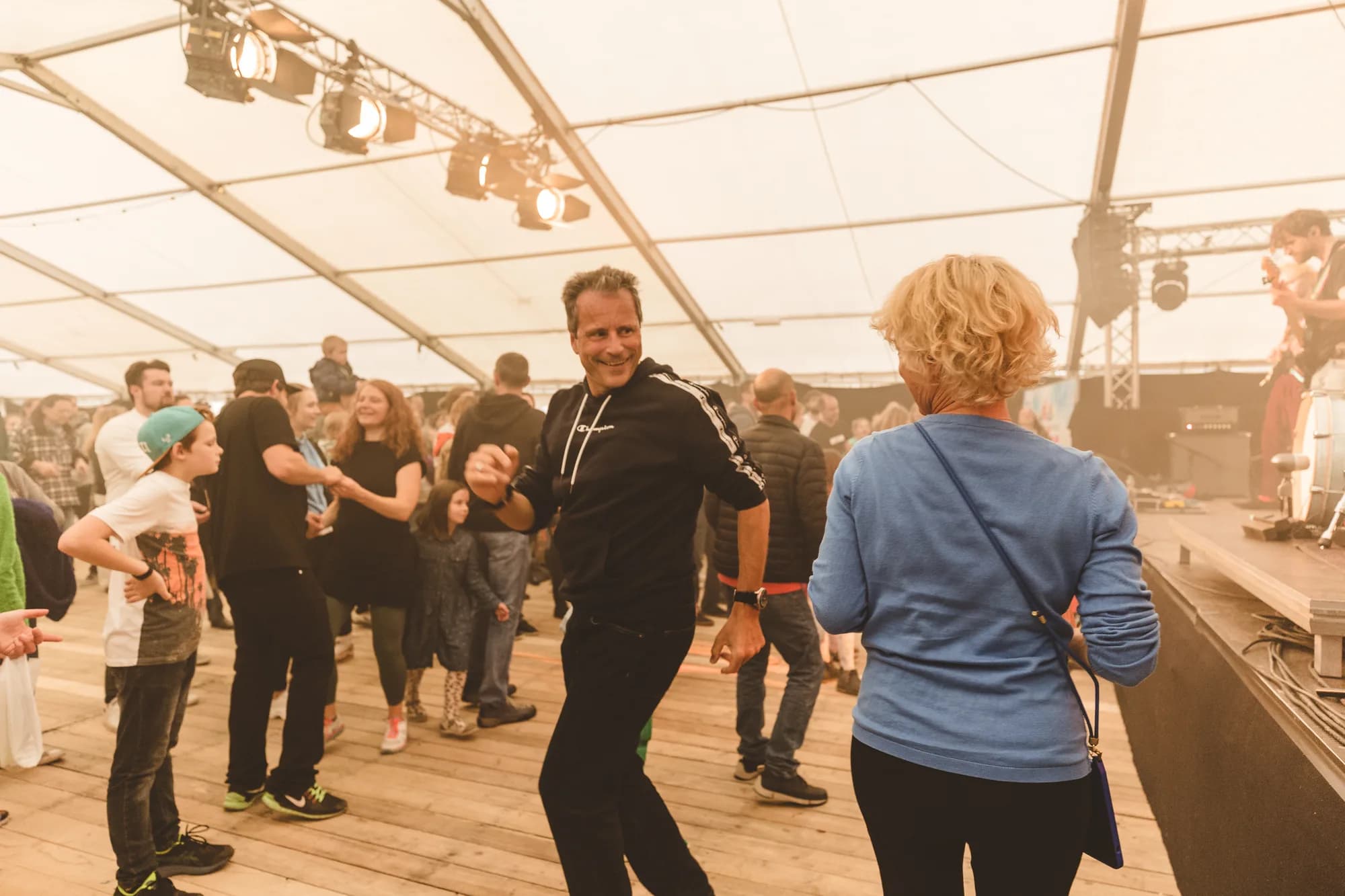 A smiling man in a black hoodie dances energetically on a wooden stage floor while a DJ performs in the background.