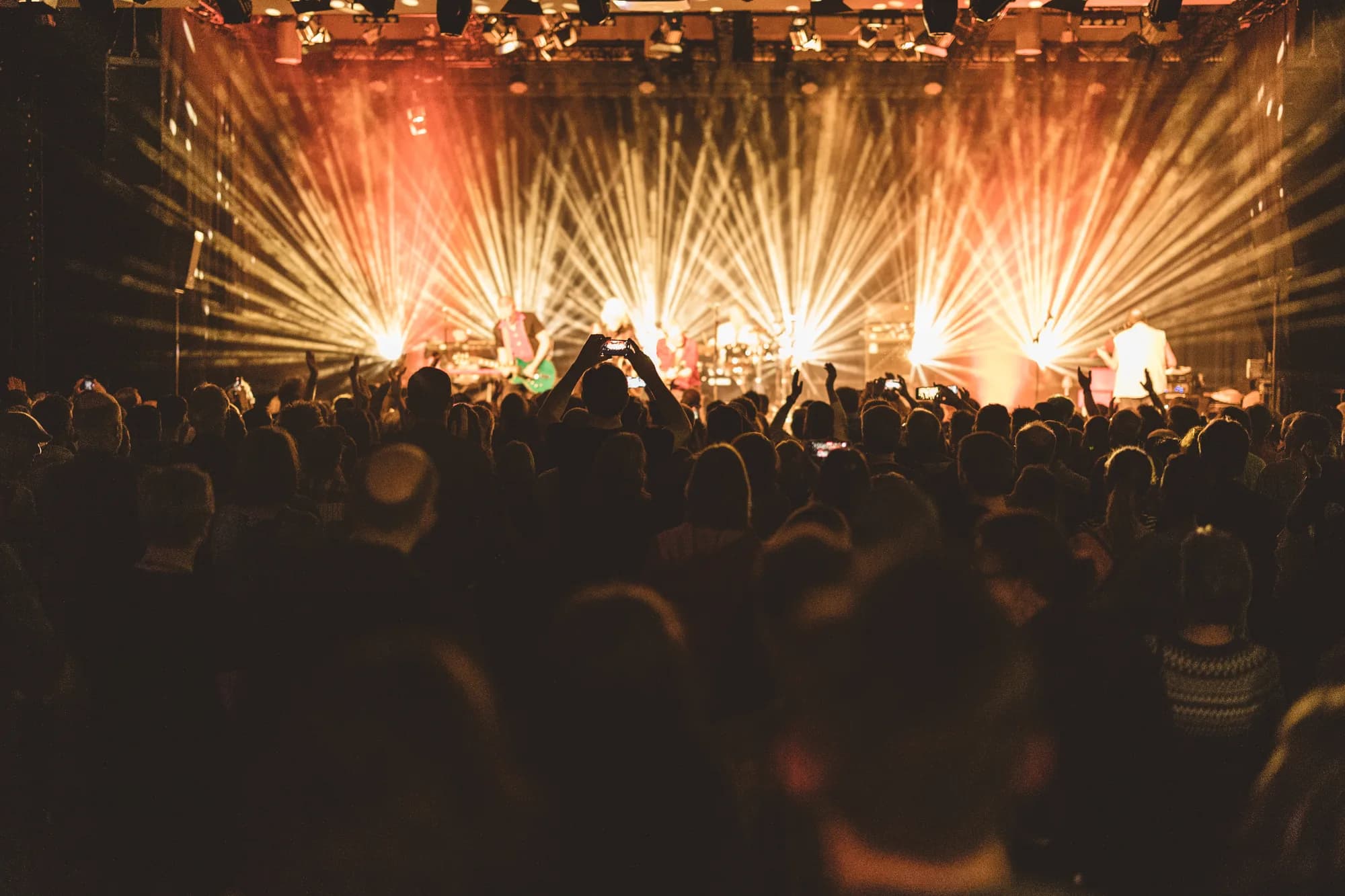 A DJ performs on stage illuminated by a massive fan of orange and white light beams over a dense crowd.