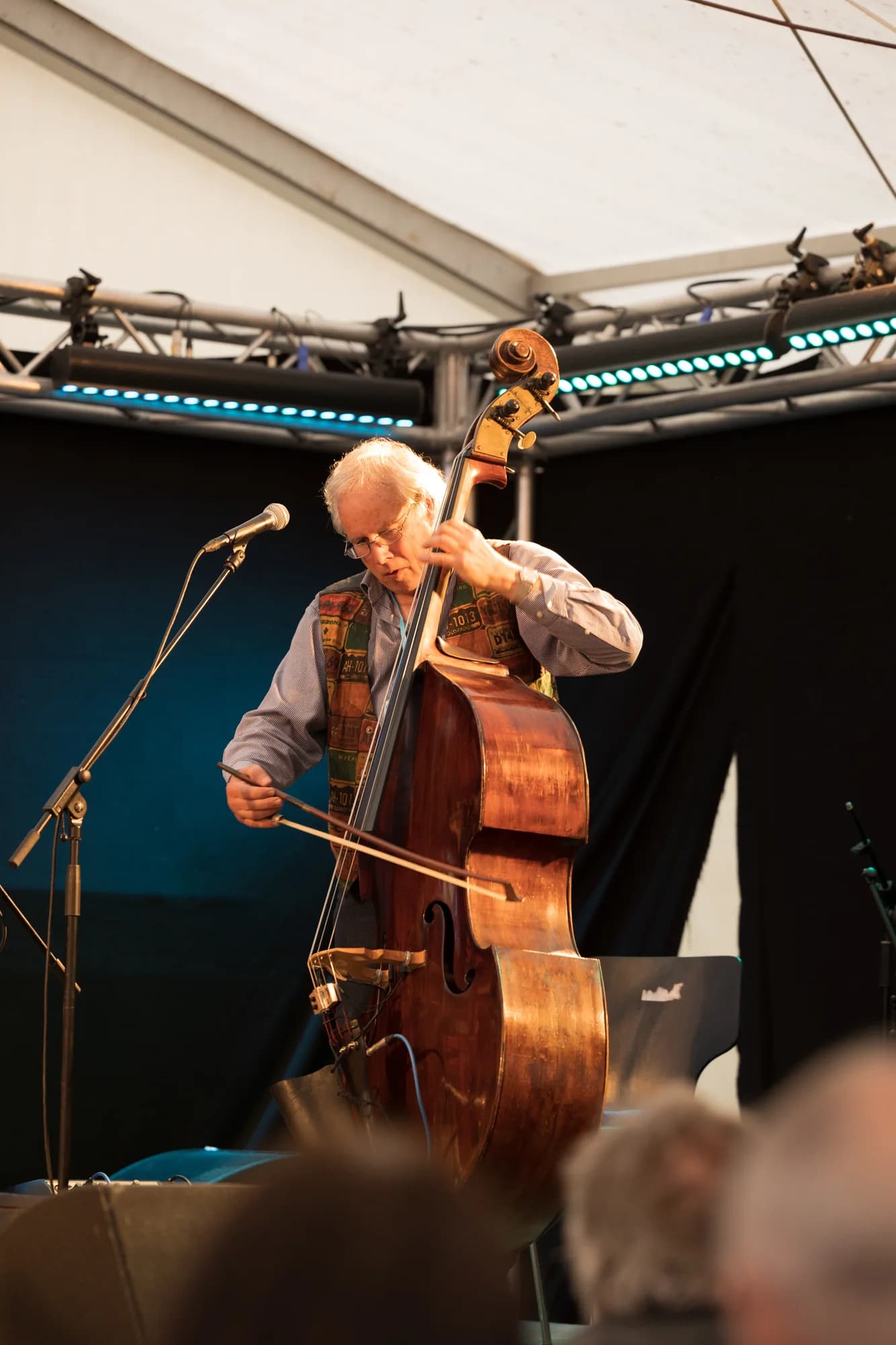 An older musician with white hair plays a wooden double bass with a bow on a stage with blue lighting.