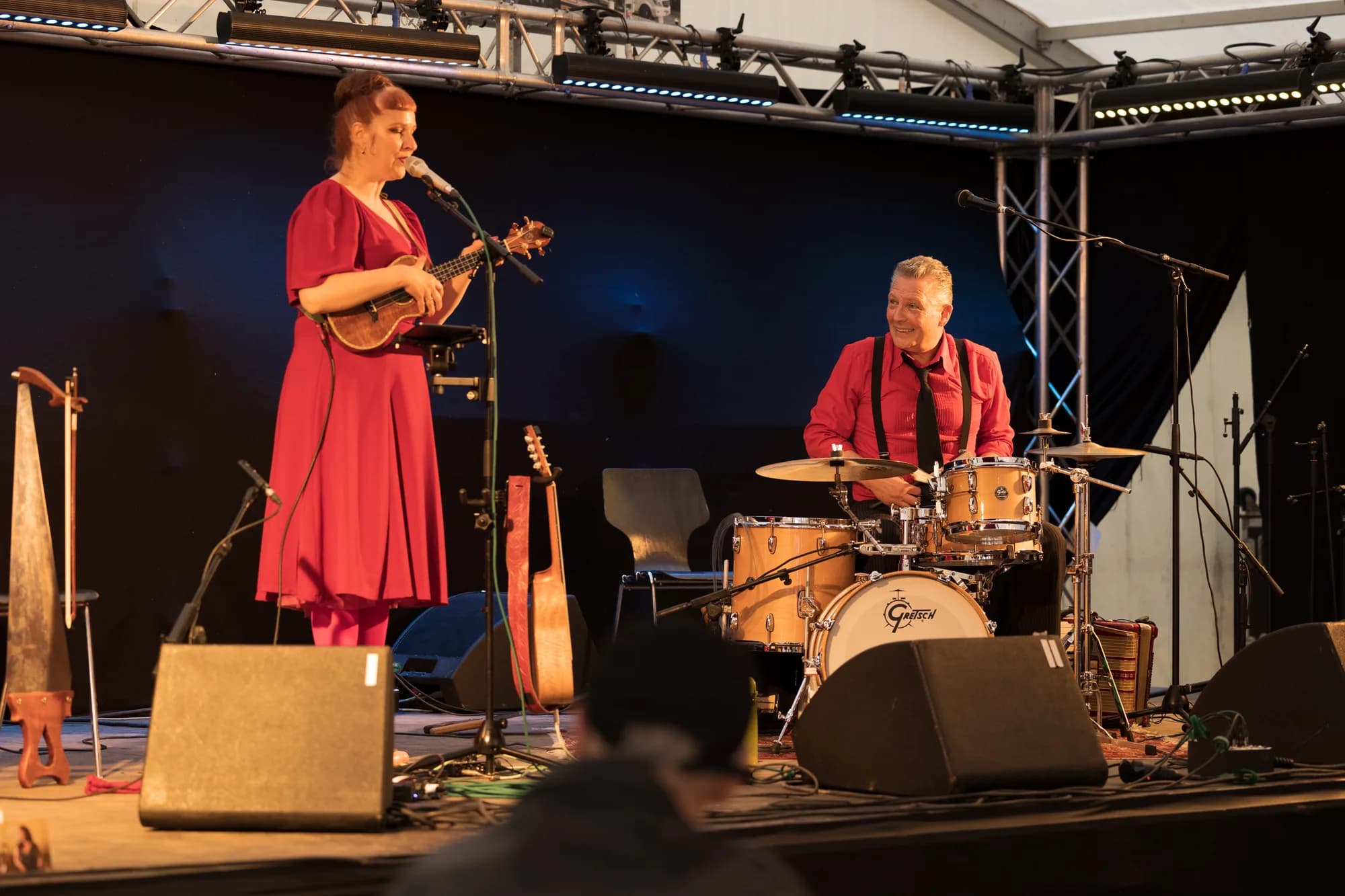 A female vocalist in a red dress plays a ukulele while a male drummer in a red shirt plays beside her on an outdoor stage.