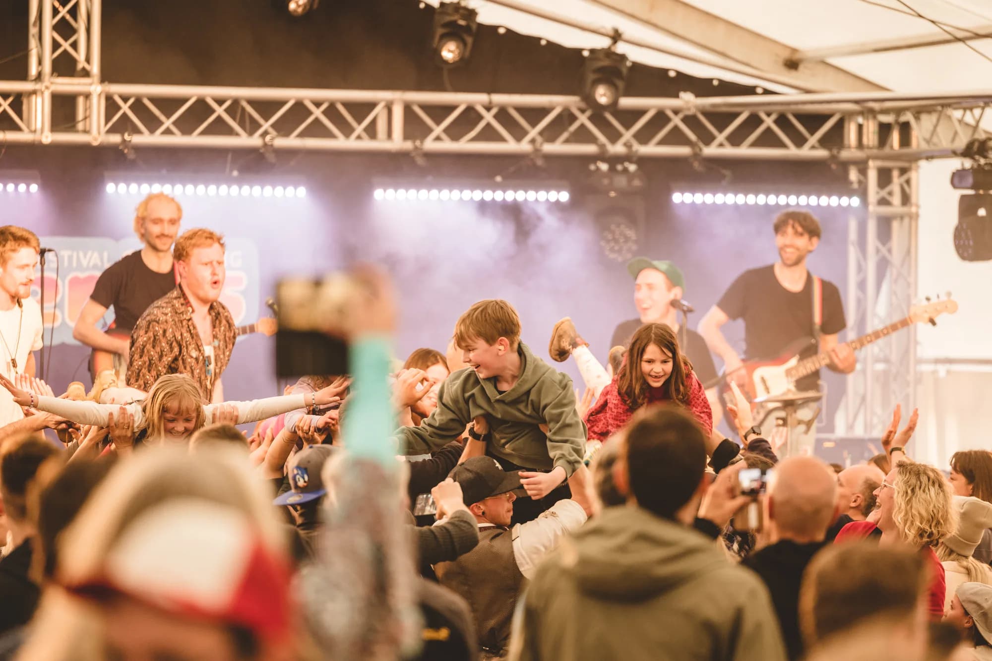A rock band performs on stage while a crowd surfer is lifted high above the audience in a packed tent.