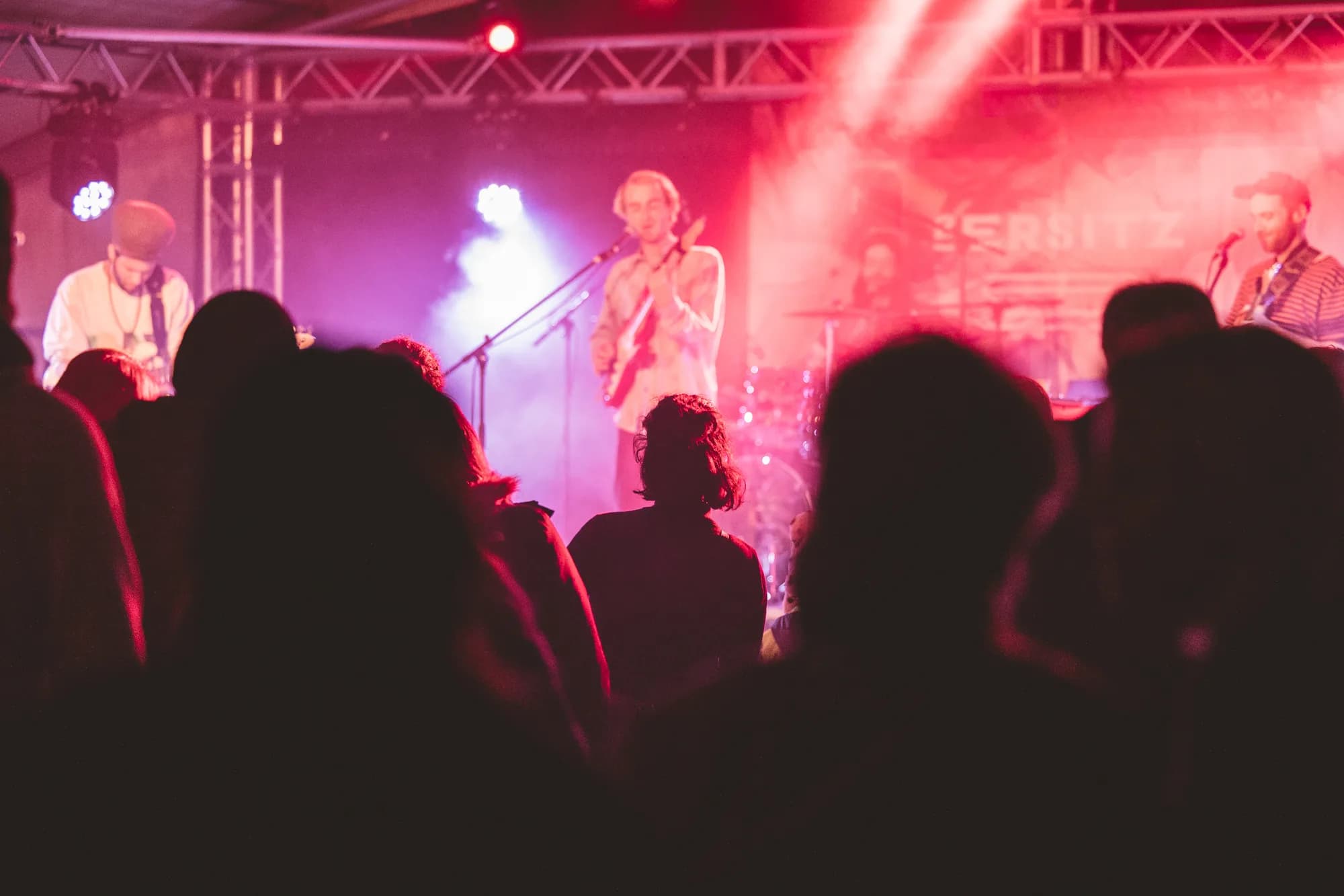 A rock band performs on stage under red and purple lights with a banner reading ERSITZ behind them.