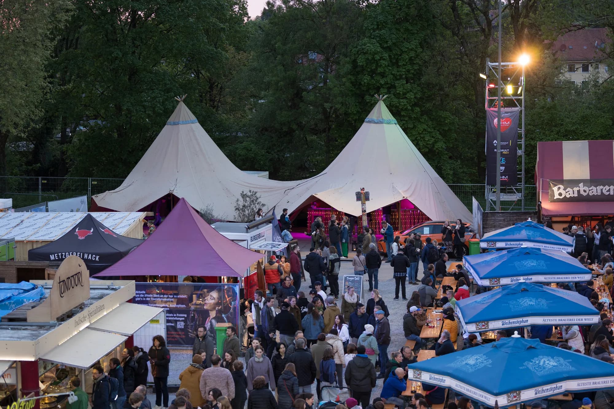 A wide shot of a busy festival market area at dusk with two large white tipis and crowds of people walking between food stalls and blue umbrellas.