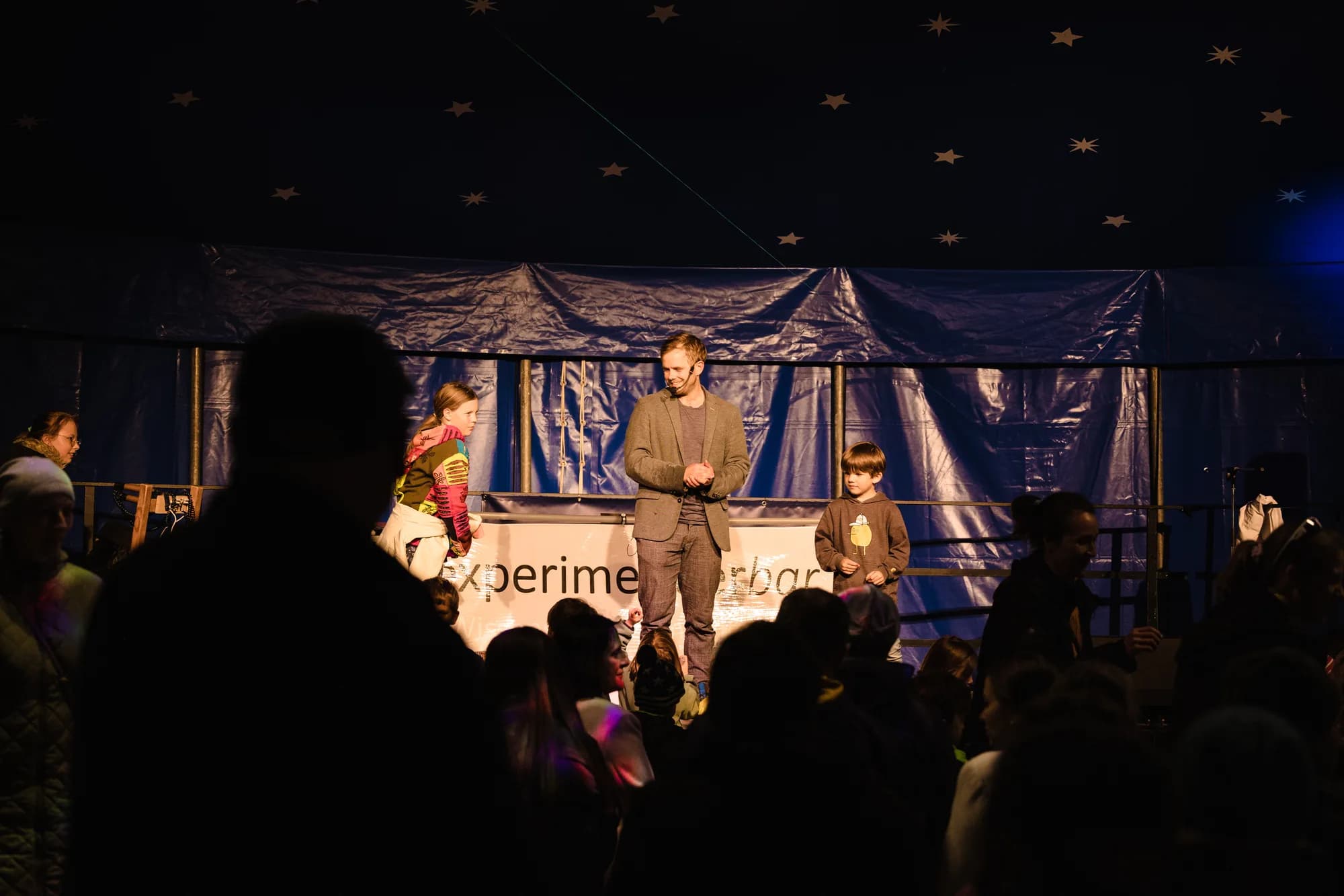 A man and two children perform on a stage under a starry tent ceiling with an audience watching.