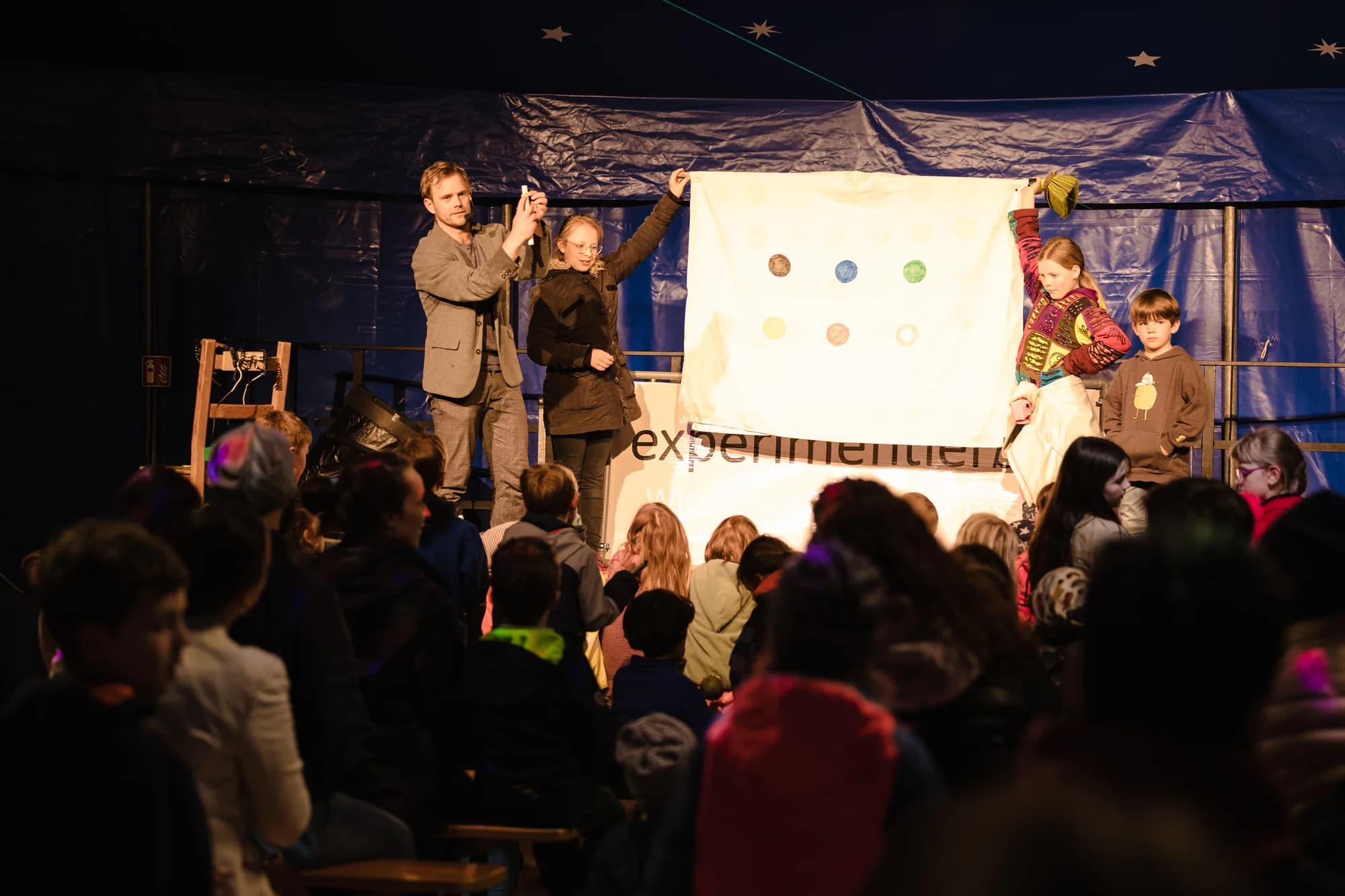 Performers and children on stage holding a white banner with colorful dots during a children's theater show.