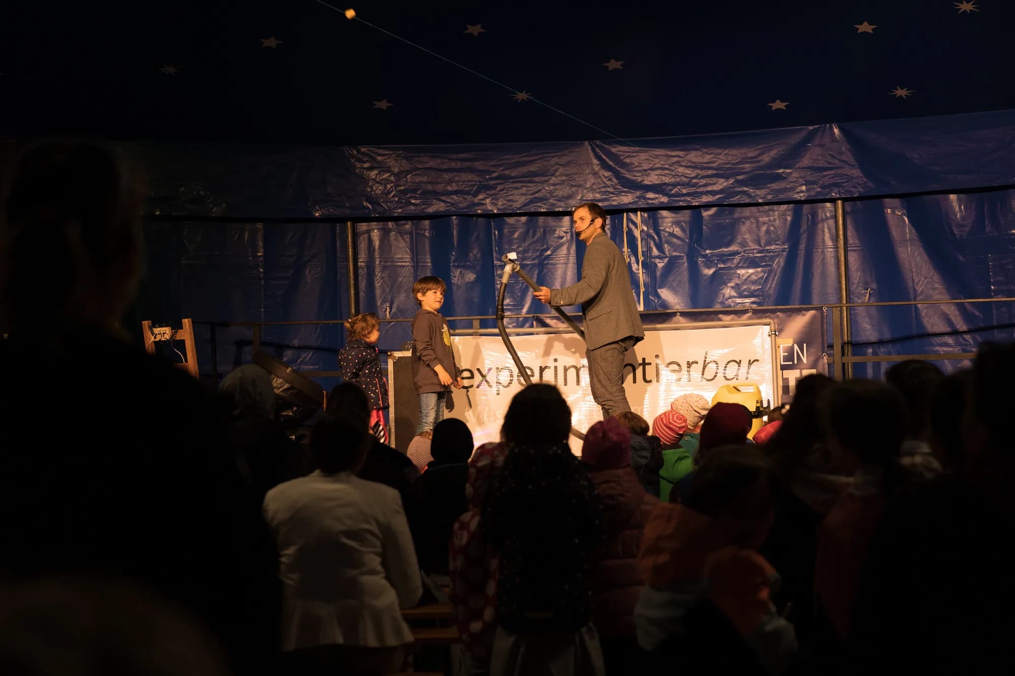 A performer and two children on stage with a large hose prop during an evening show under a starry tent.