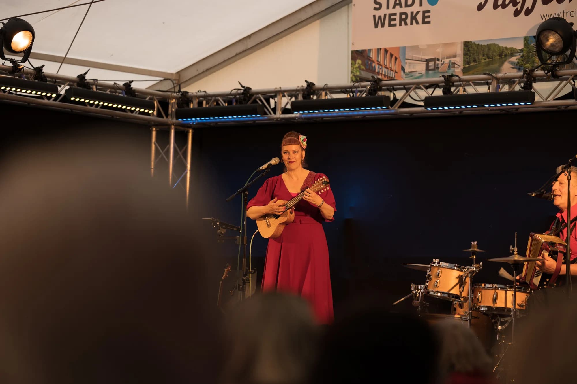 A female musician in a long red dress plays a ukulele on stage under warm spotlights.