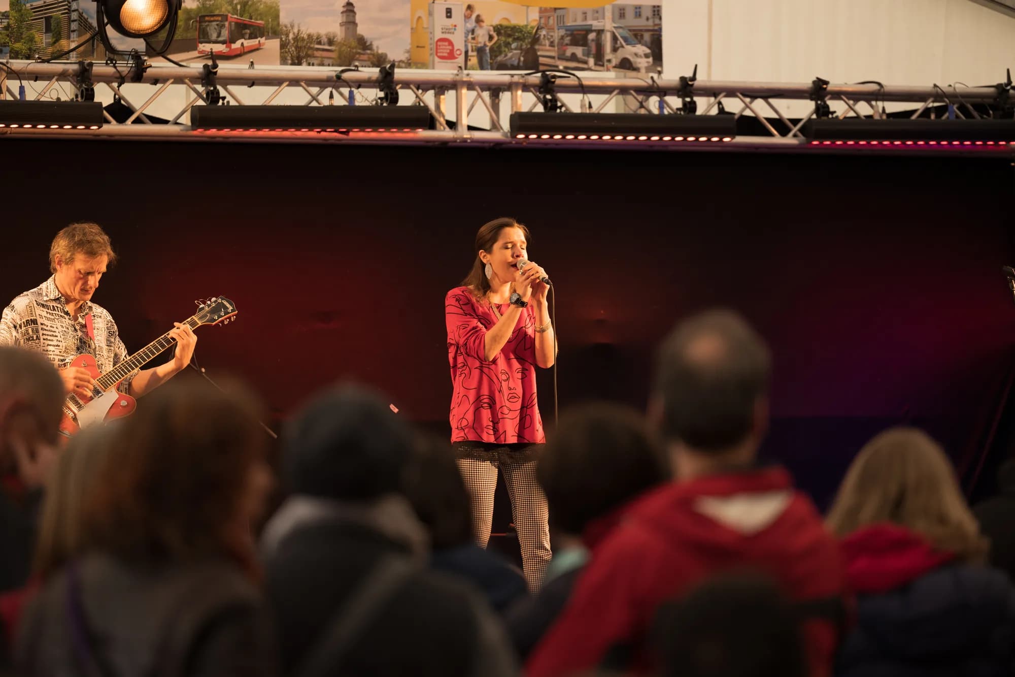 A female singer in a pink shirt performs with a guitarist on an outdoor stage in front of an audience.