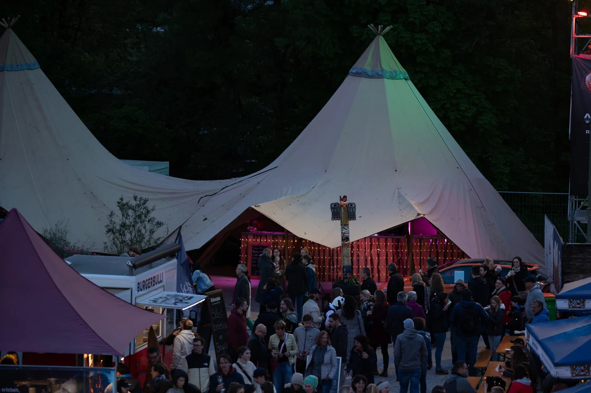 A view of large white teepee tents illuminated by pink and green lights at the Uferlos Festival with a crowd gathered in front.
