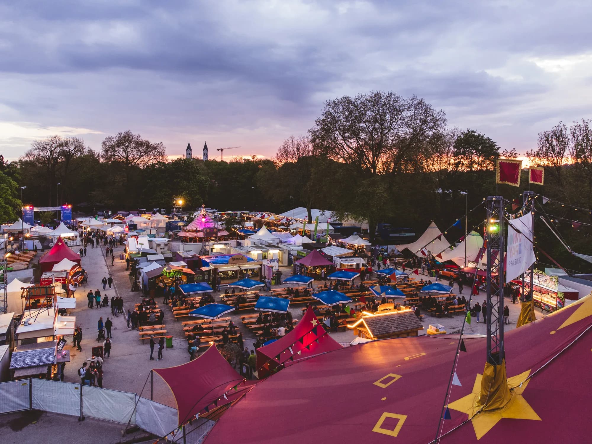 Aerial view of the Uferlos festival market at dusk with red tents and people dining under blue umbrellas.