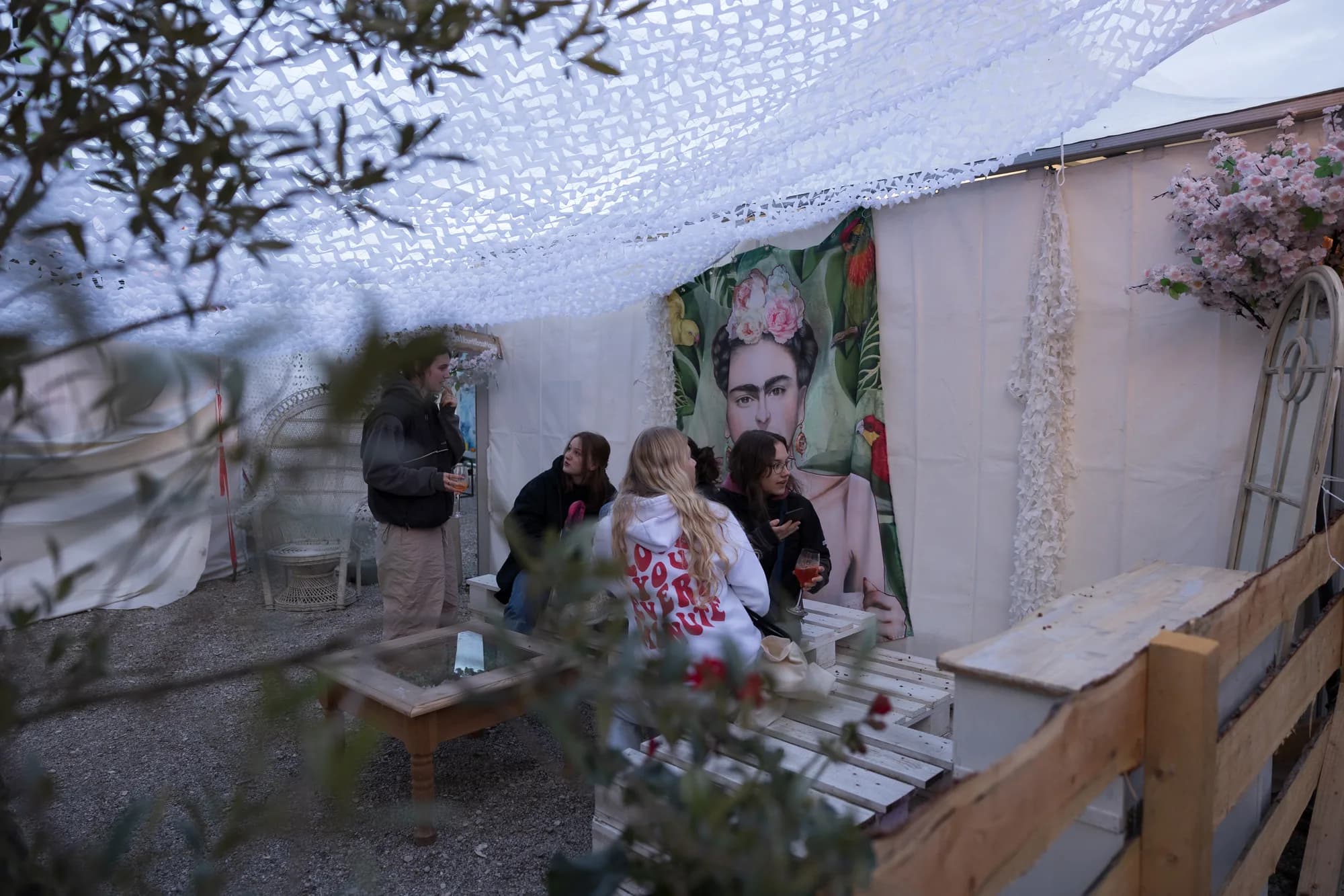Festivalgoers relax on wooden pallets inside a white tent decorated with a large Frida Kahlo poster and hanging white netting.