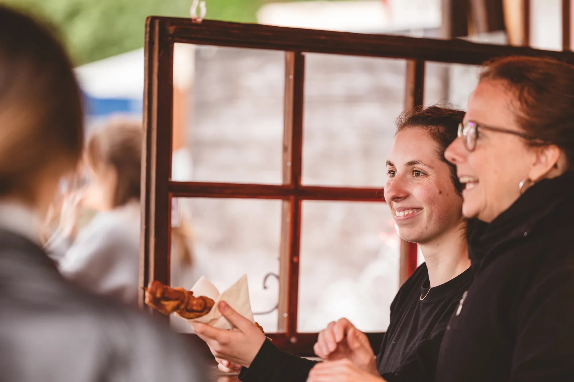 Two smiling women sit at a wooden bar counter, one holding a fried pastry while chatting with a server.