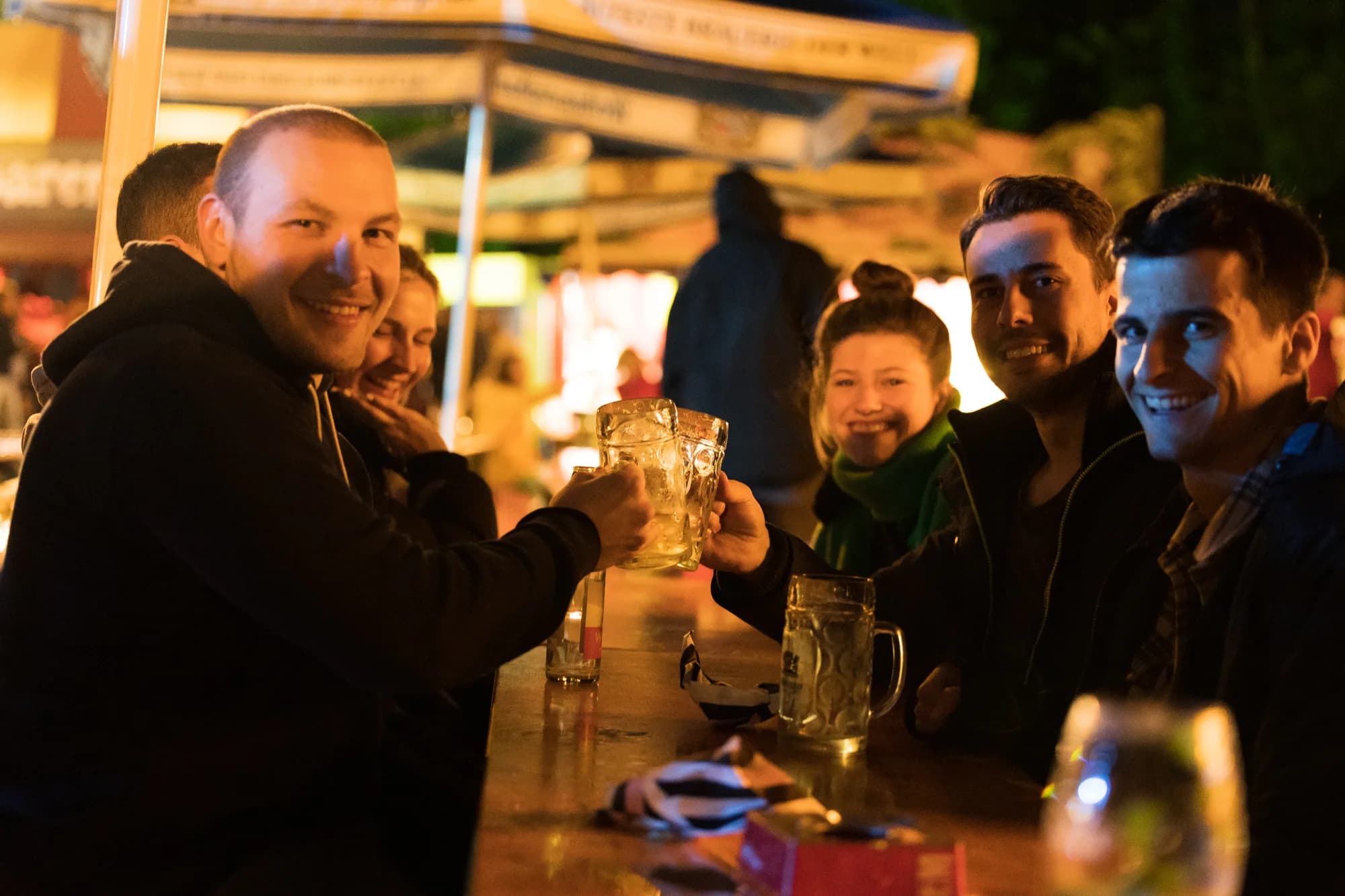 A group of smiling friends clinking large glass beer mugs together at a wooden bar counter at night.