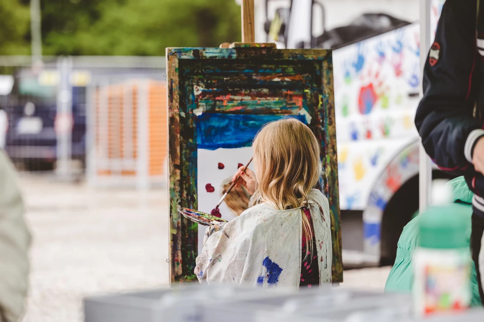 A young girl with blonde hair paints on a canvas at an easel during a children's workshop.