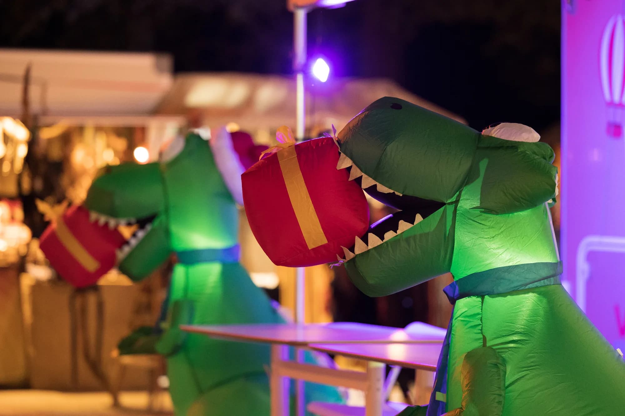 Two green inflatable T-Rex costumes holding red gift boxes illuminated by purple stage lights at night.