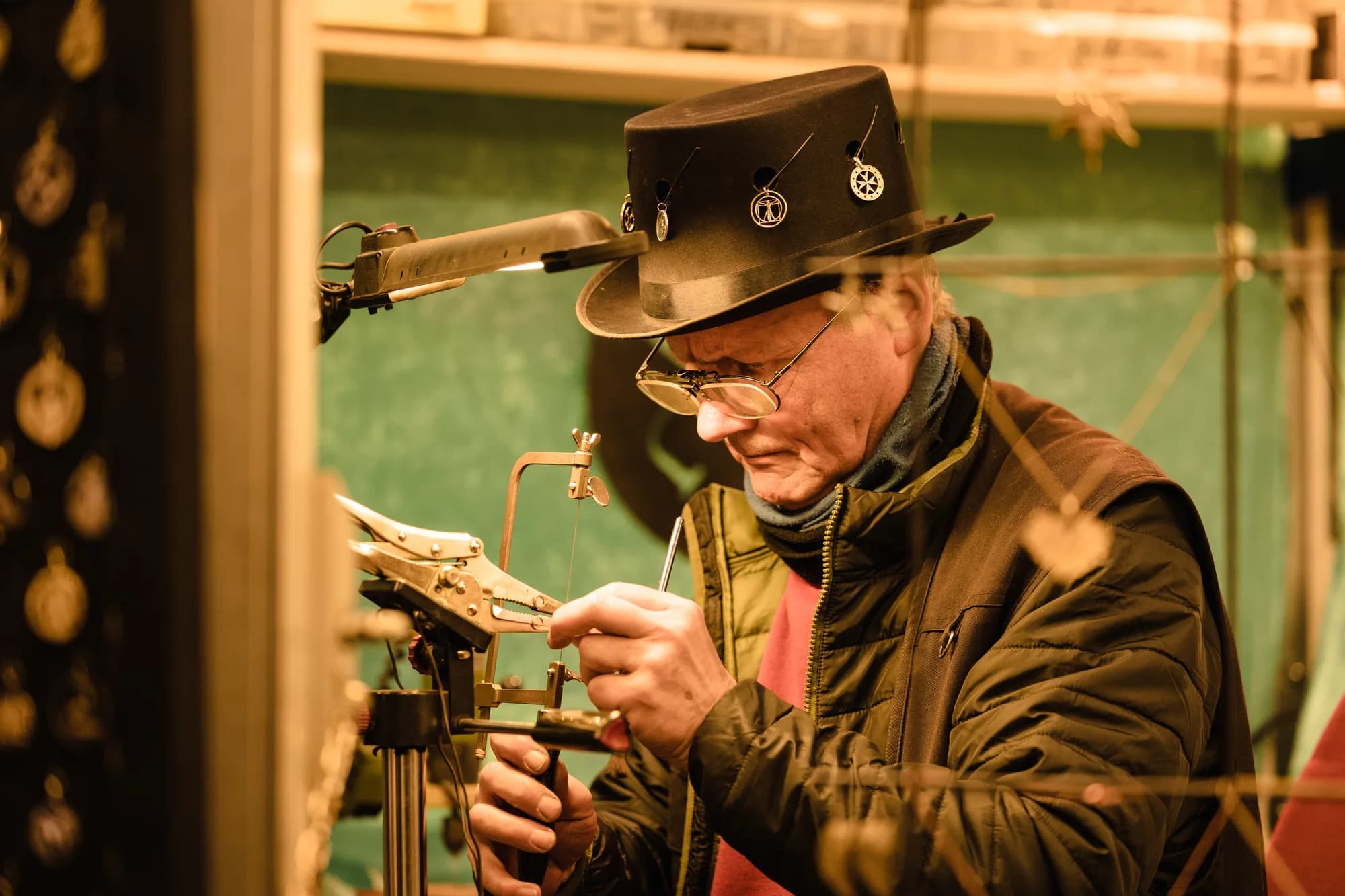 A craftsman wearing a hat and glasses uses a jeweler's saw to cut metal at a market stall.