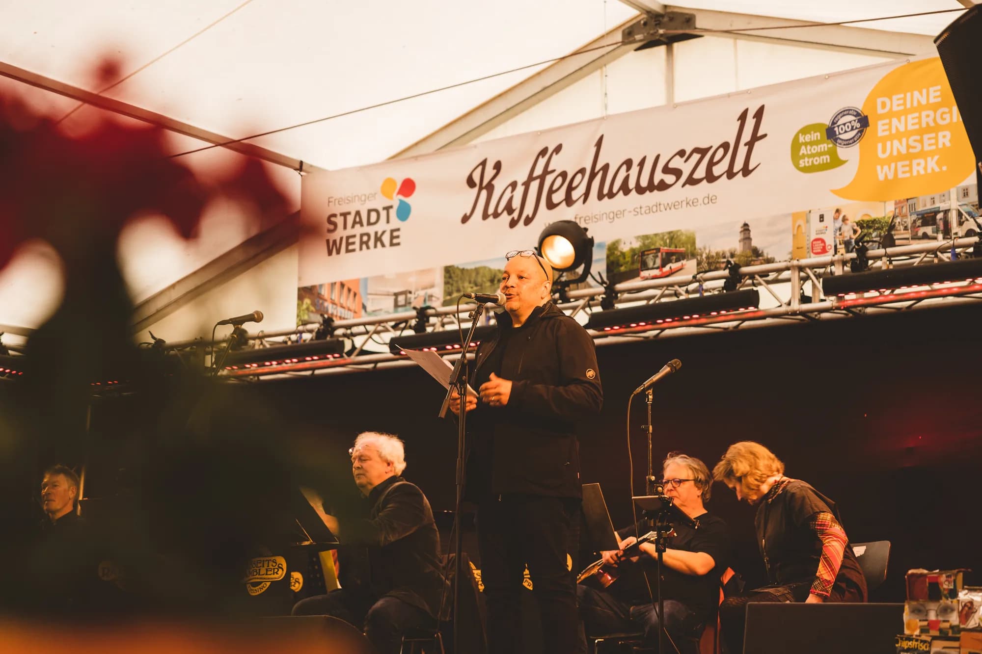 A band performs on the Kaffeehauszelt stage with a singer holding a microphone and musicians playing instruments.