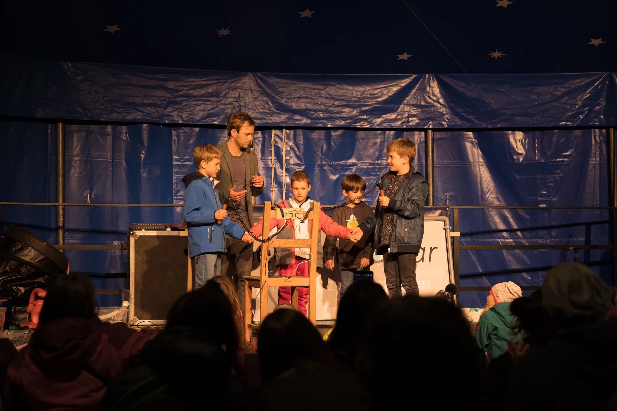 A male performer and four young children stand on a wooden stage holding hands in front of a blue curtain backdrop.