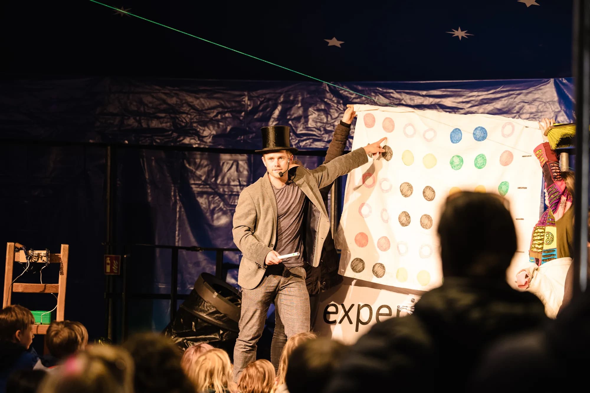 A male magician in a top hat performs a magic trick with a colorful board for an audience of children.