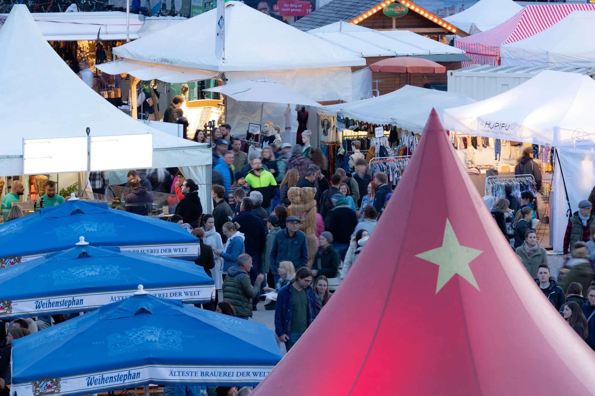 A crowded market area with white vendor tents and blue beer umbrellas, featuring a large red tent with a yellow star in the foreground.