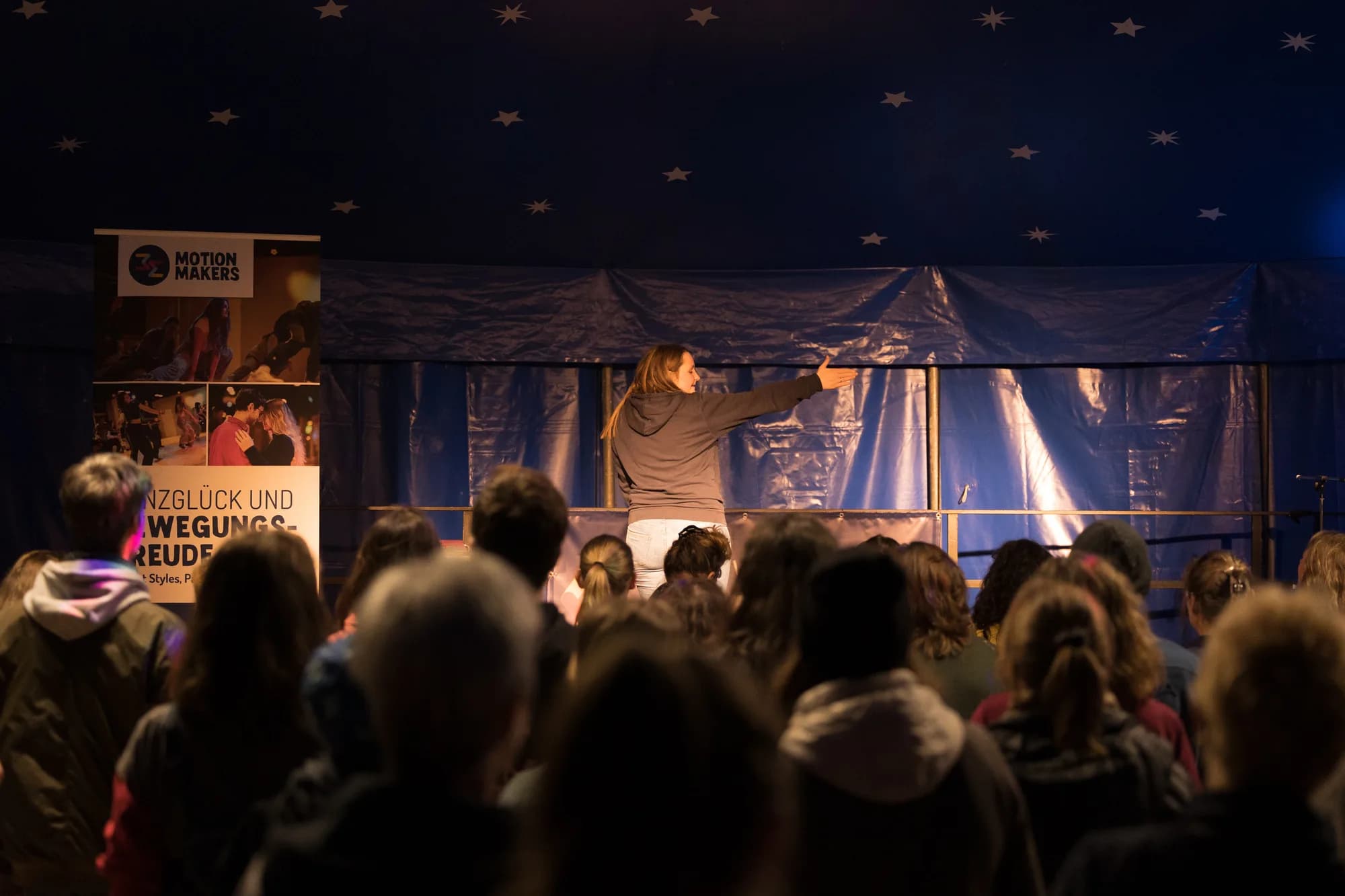 A female instructor leads a movement workshop on stage in front of a seated audience inside a dark tent.